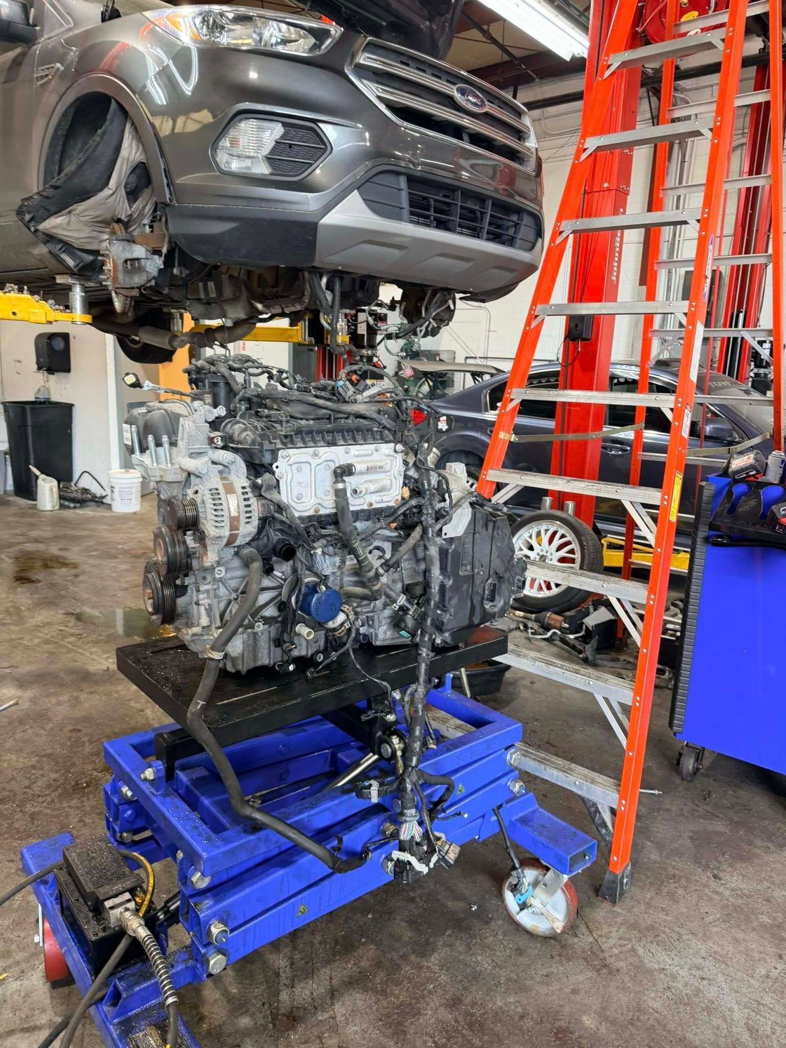 Car suspended above a car engine on a blue lift in an auto repair shop with a red ladder nearby | Best Choice Service