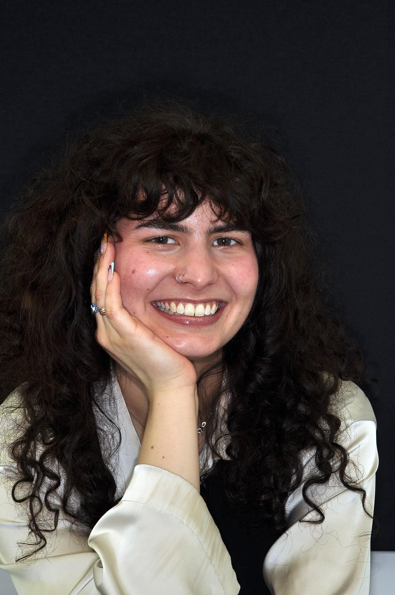 Persona sorridente con capelli scuri e ricci che appoggia il mento sulla mano, su uno sfondo scuro.