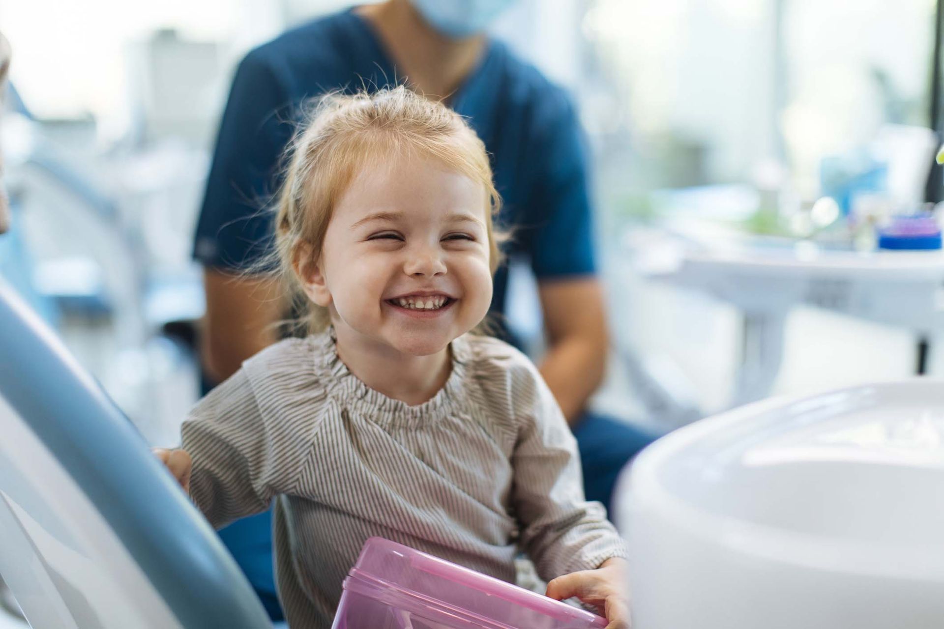 Bambina sorridente seduta sulla poltrona del dentista, con in mano una cartella rosa, e un dentista sullo sfondo.