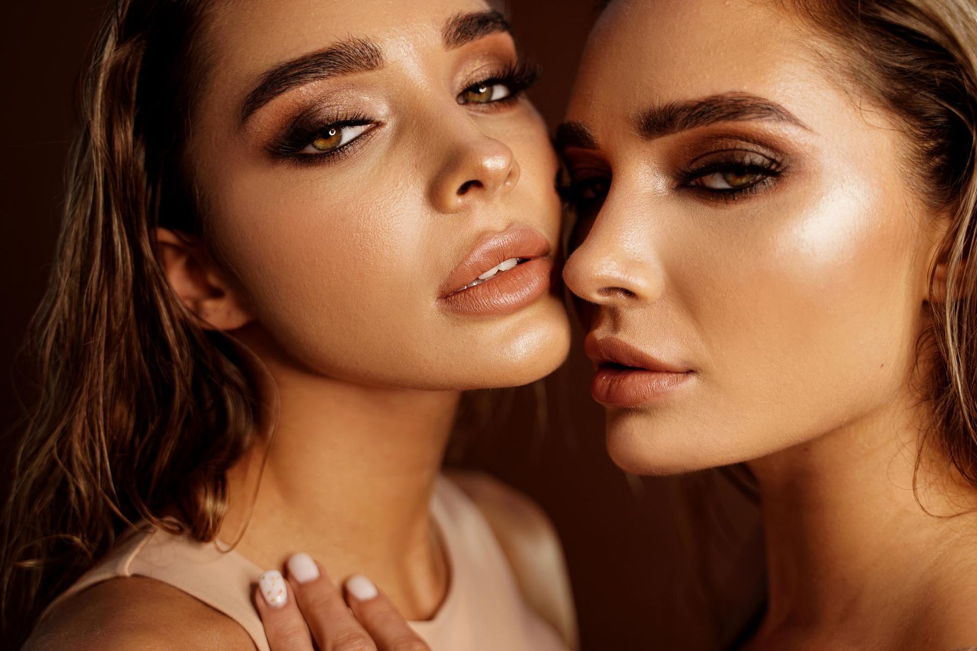 Due donne con un trucco dai toni caldi posano vicine su uno sfondo scuro.