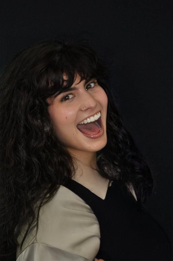 Donna dai capelli scuri e ricci, sorridente su uno sfondo scuro, indossa una maglietta nera e beige.