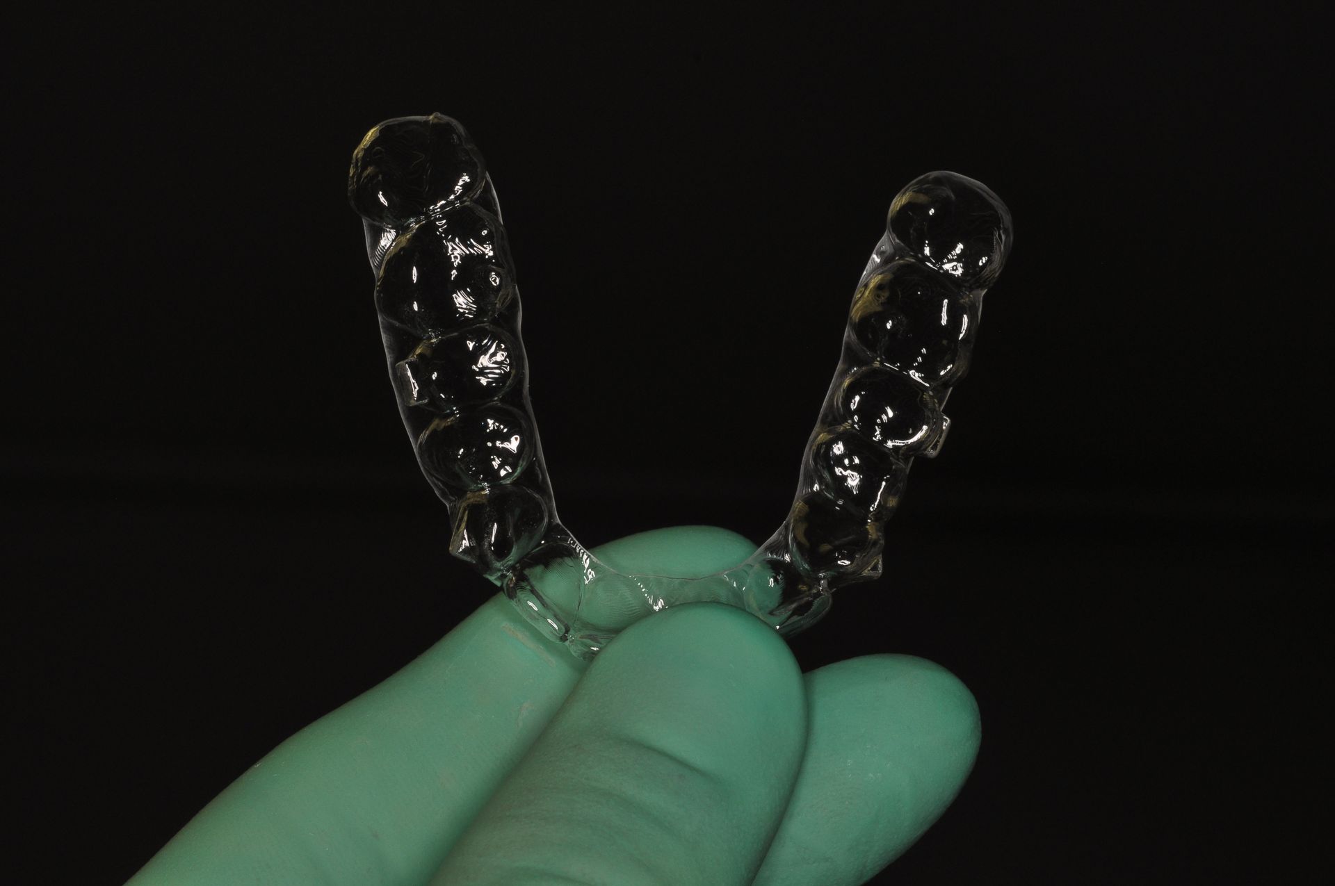 Mano guantata che regge due allineatori dentali trasparenti su sfondo nero.