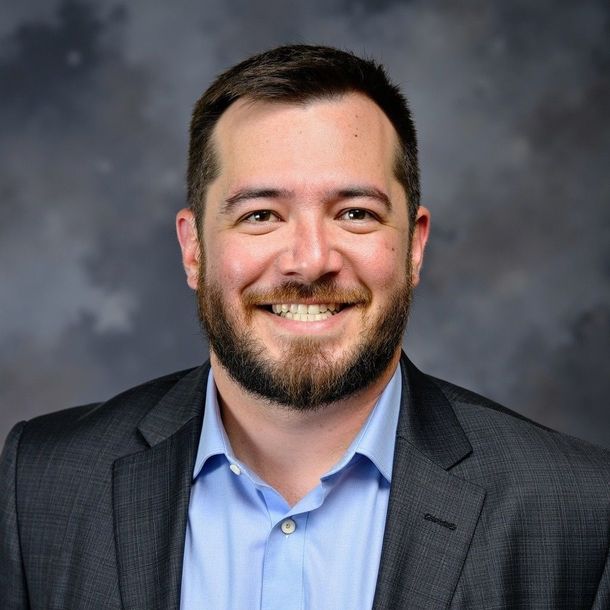 Man in a dark suit jacket, white shirt, smiling. Gray background.