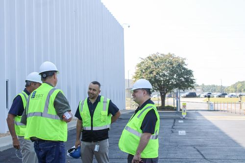 Four people in safety vests and hard hats stand outside a building, conversing near a paved area.