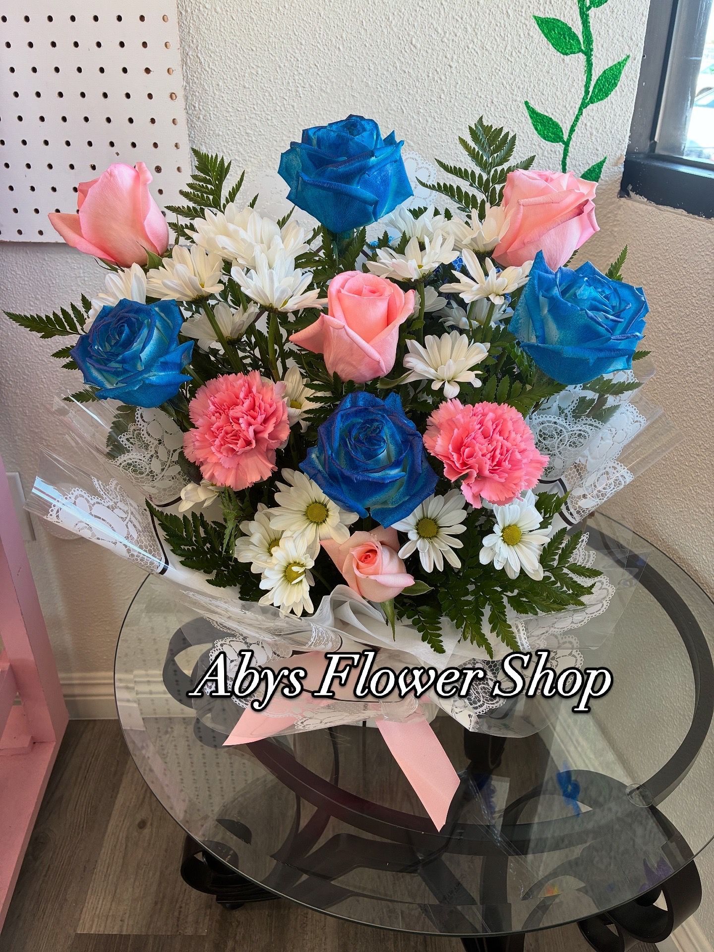 A floral arrangement of blue roses, pink roses, and white daisies in a glass vase on a glass table.