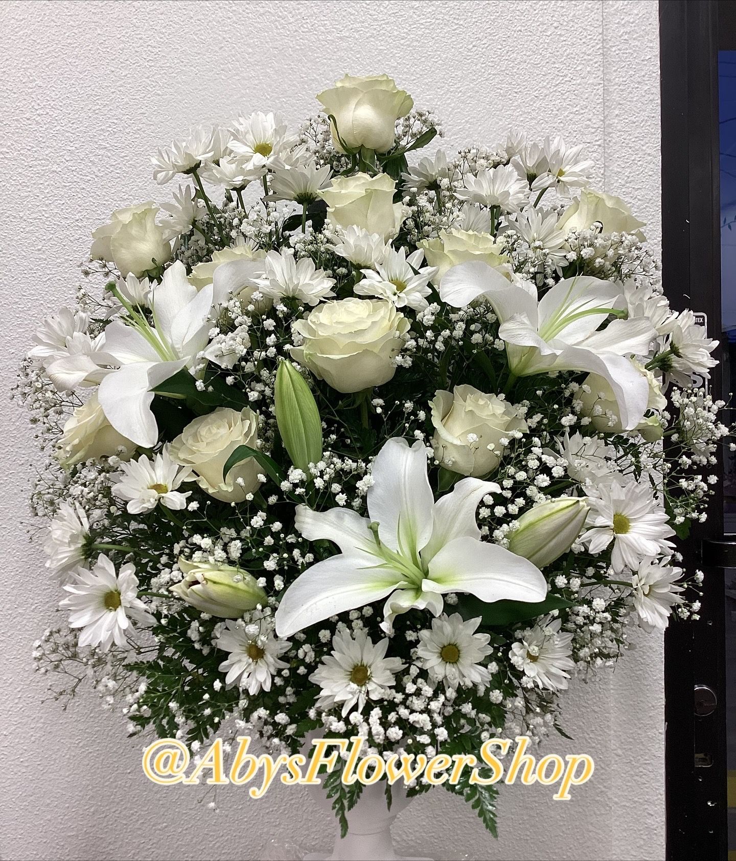 A round arrangement of white lilies, roses, and daisies with baby's breath, set against a plain light wall.