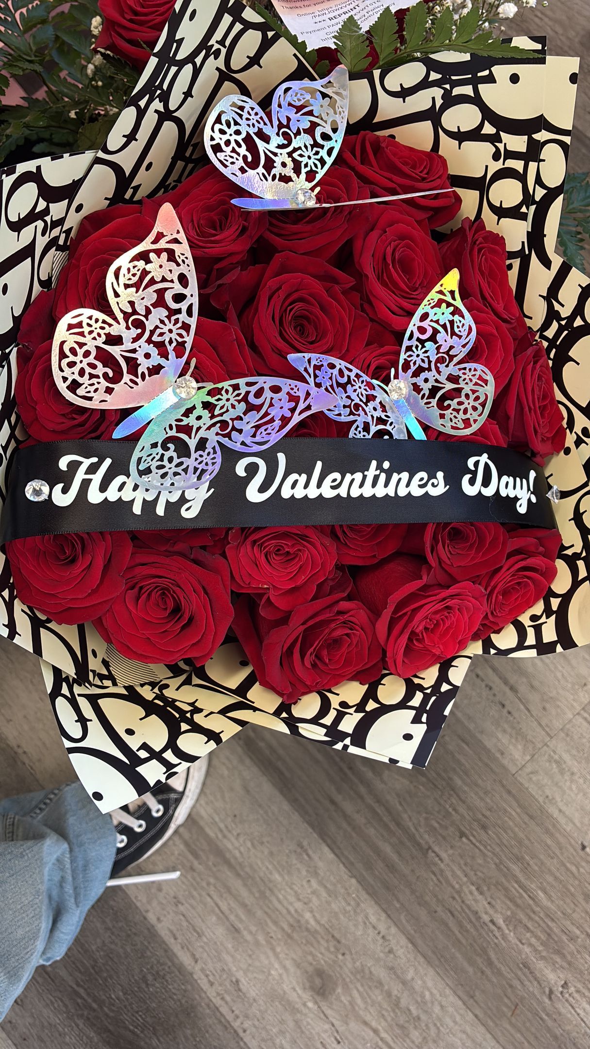 A bouquet of red roses wrapped in patterned paper, decorated with silver butterfly ornaments and a Valentine's Day ribbon.