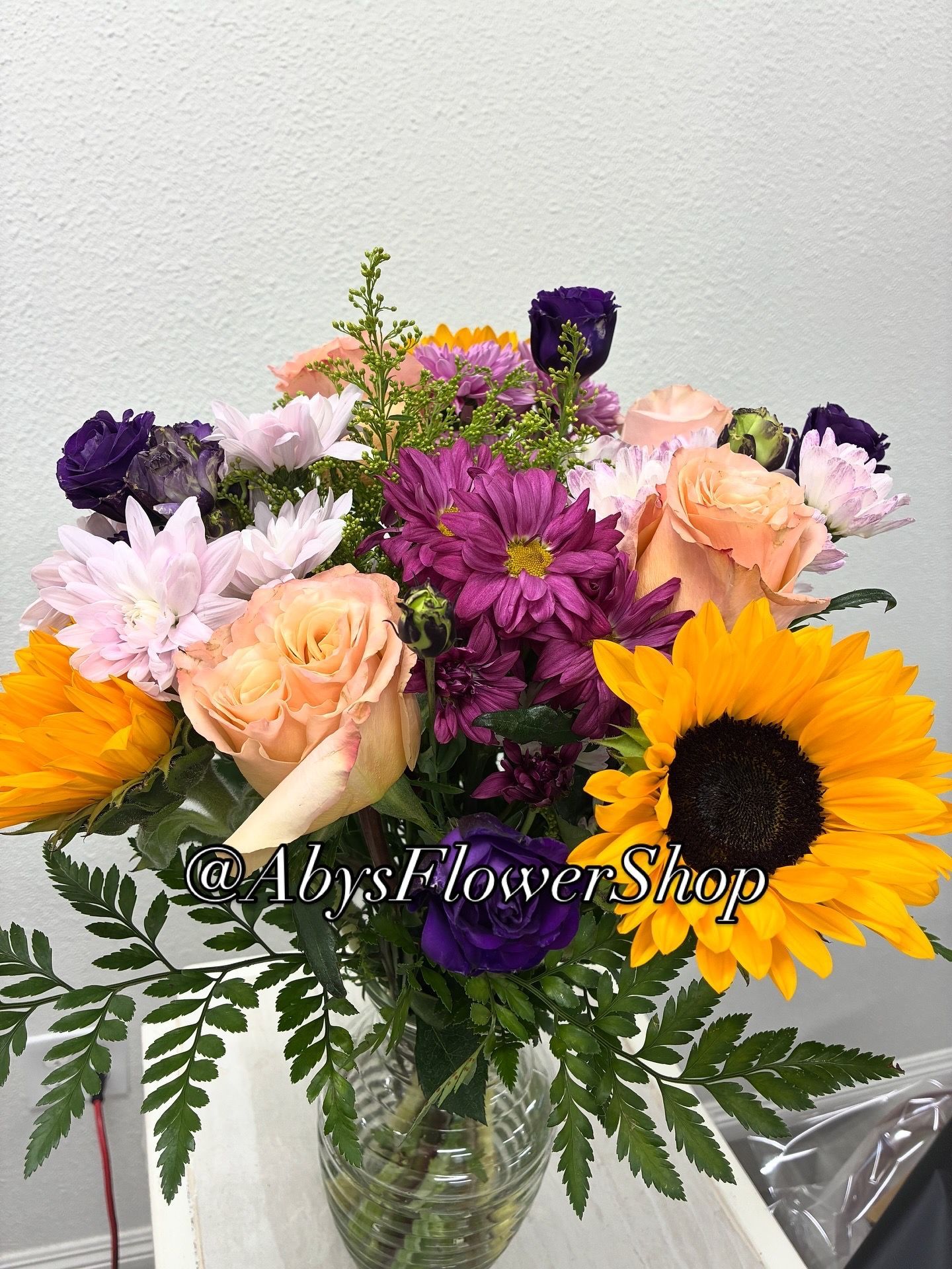 A vibrant floral bouquet in a glass vase featuring sunflowers, peach roses, purple daisies, and green foliage.