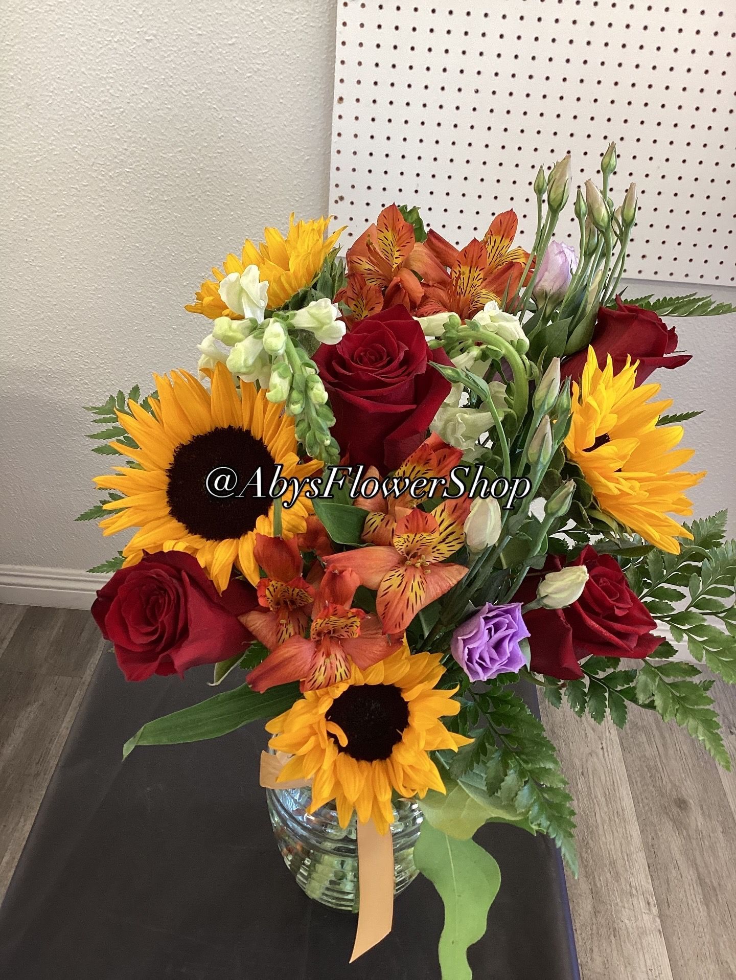 A vibrant bouquet of sunflowers, deep red roses, and orange alstroemeria in a clear glass vase against a pegboard wall.