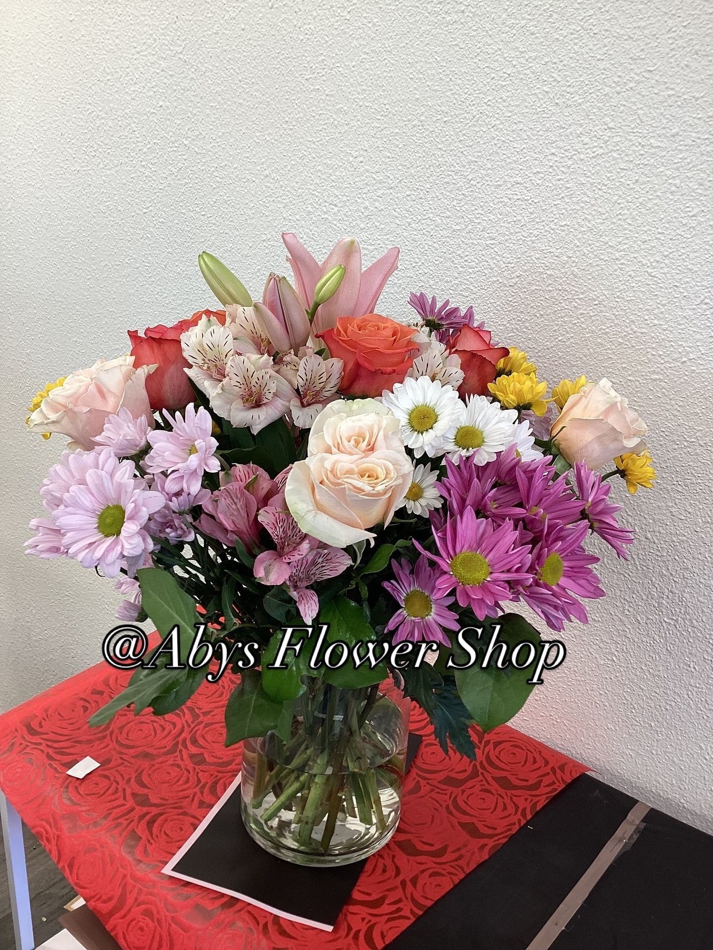 A colorful bouquet of lilies, roses, and daisies in a glass vase on a red table with a floral shop logo.