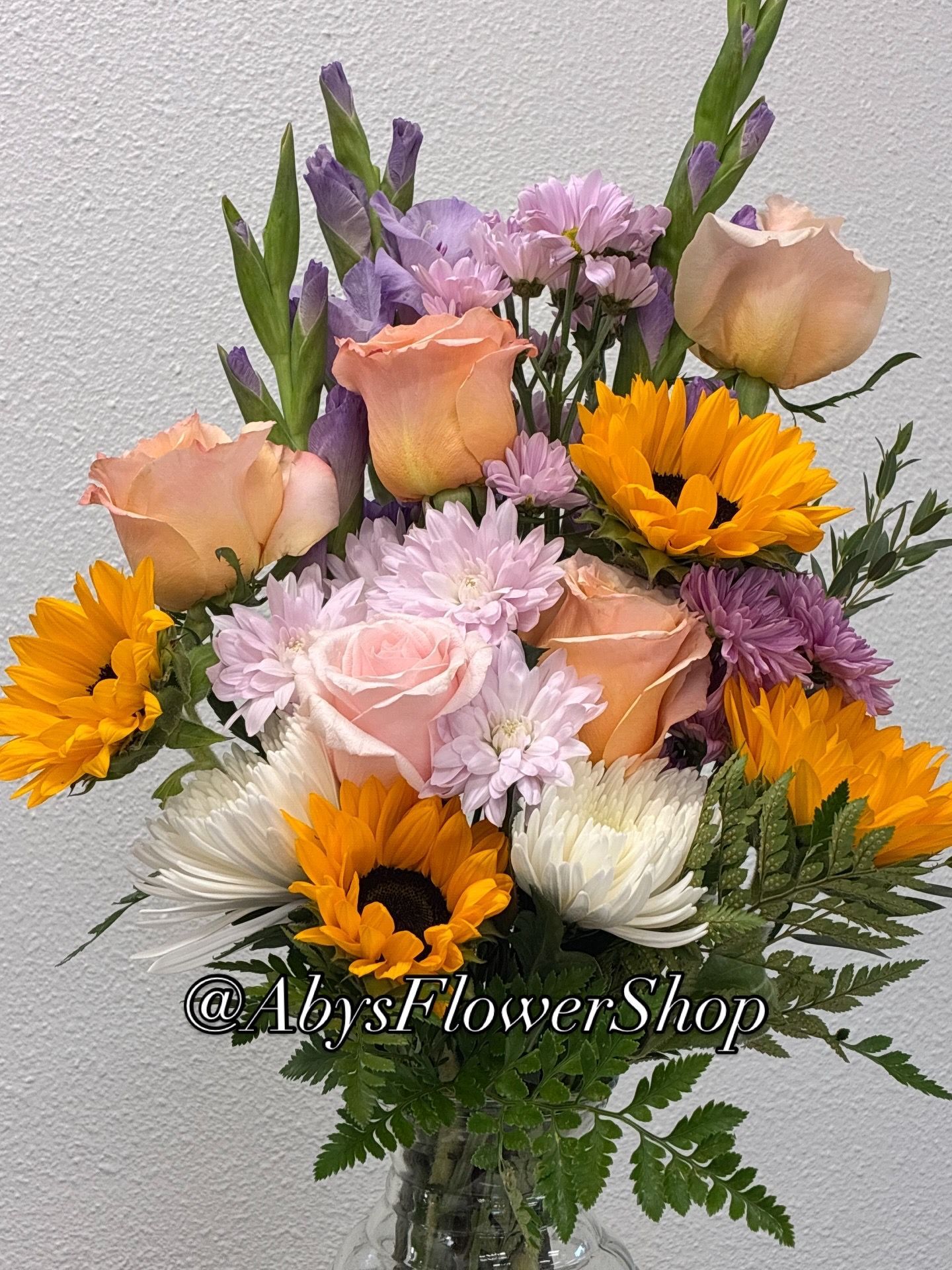 A bouquet featuring yellow sunflowers, peach roses, purple gladiolus, and white chrysanthemums with green foliage.