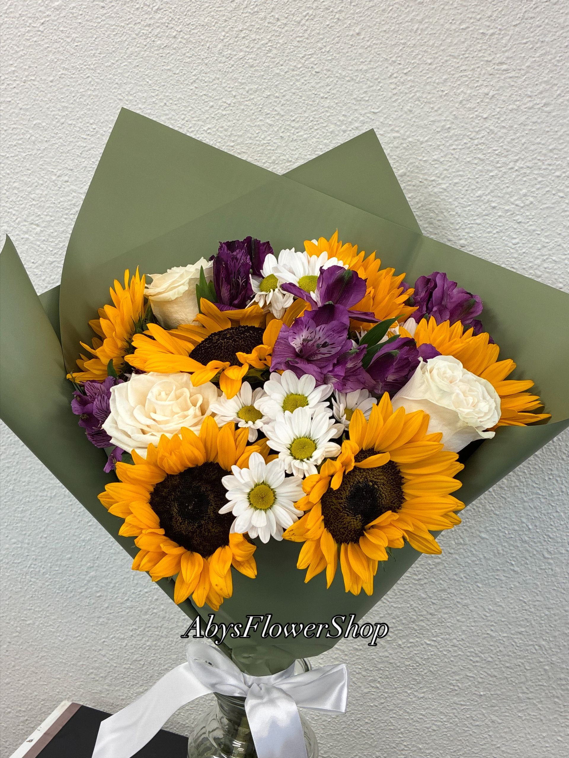 A vibrant bouquet of yellow sunflowers, white roses, purple alstroemeria, and daisies wrapped in olive-green paper.