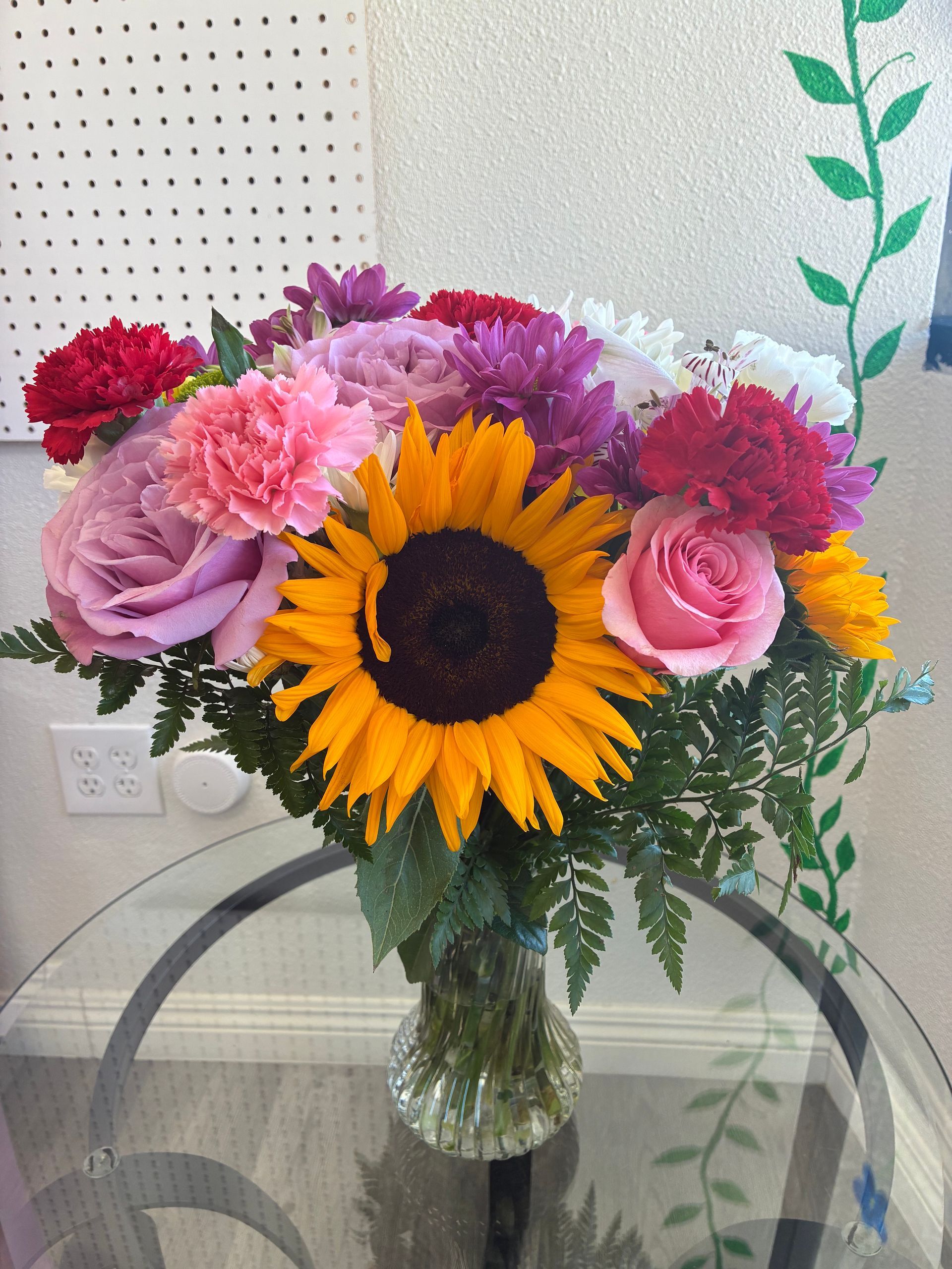 A glass vase filled with a colorful bouquet of sunflowers, pink and red carnations, and purple roses on a glass table.