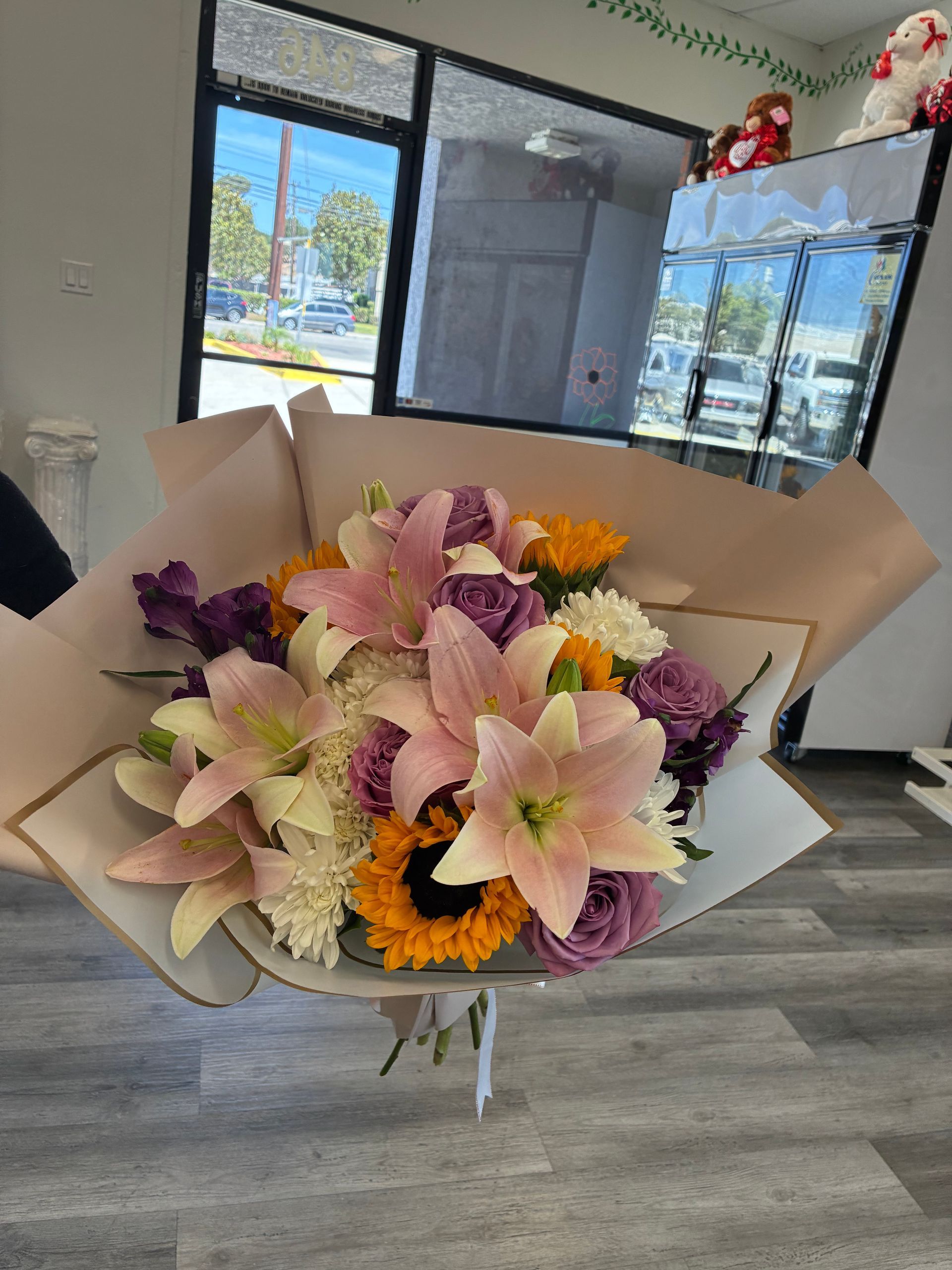 A bouquet of light pink lilies, purple roses, and sunflowers wrapped in tan paper, held indoors.