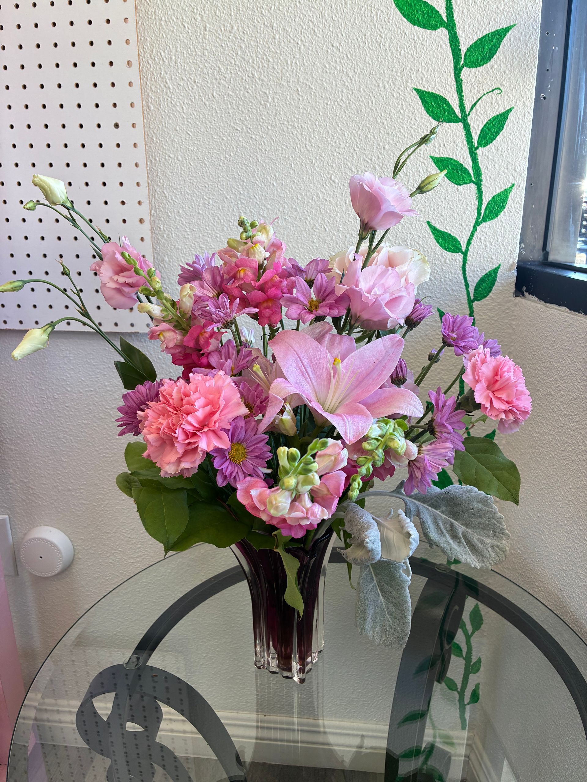 A vibrant floral arrangement in a glass vase, featuring pink lilies, carnations, and snapdragons, sitting on a glass table.