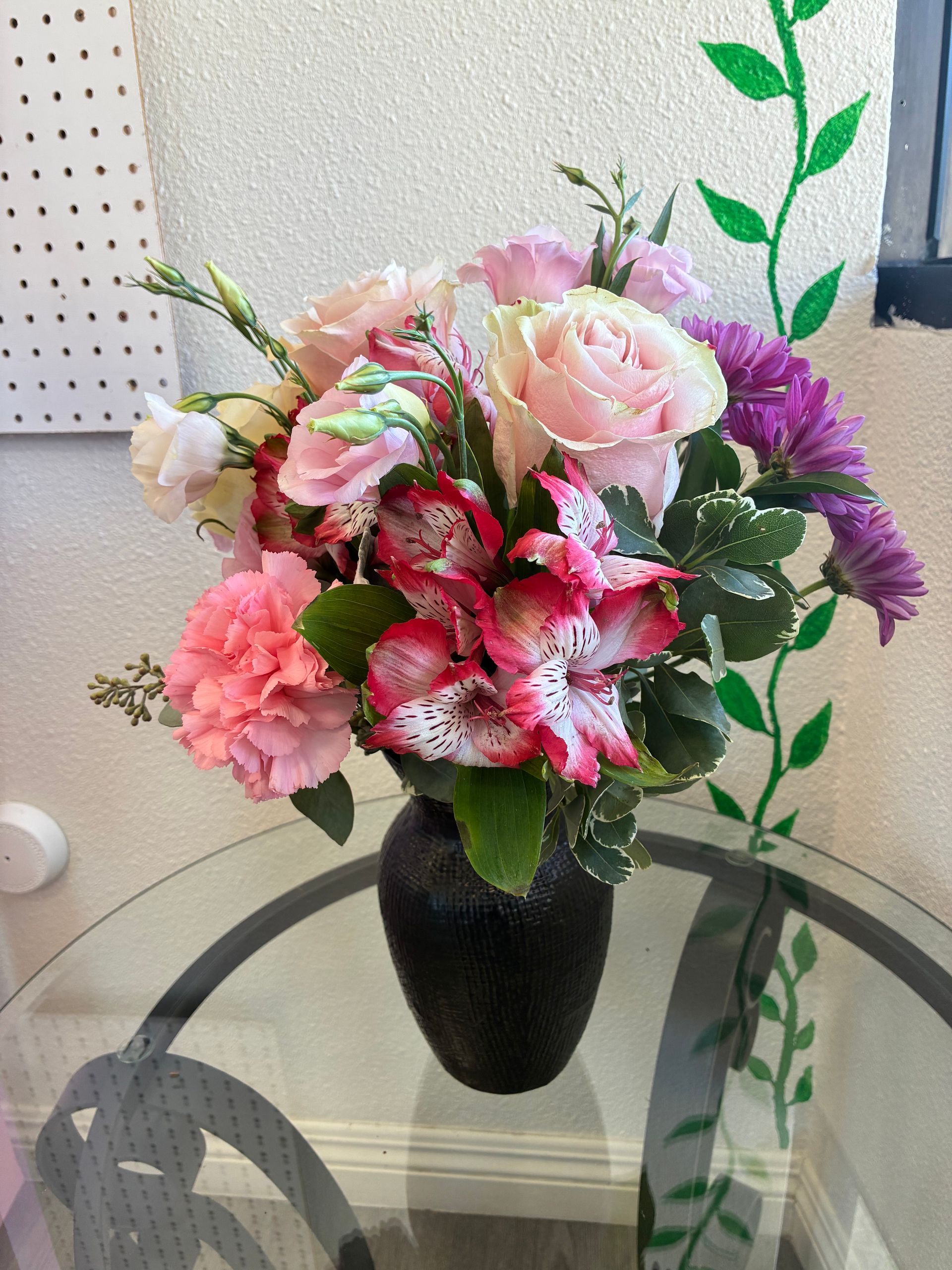 A bouquet of pink roses, spotted alstroemeria, and purple flowers in a dark vase on a glass table.
