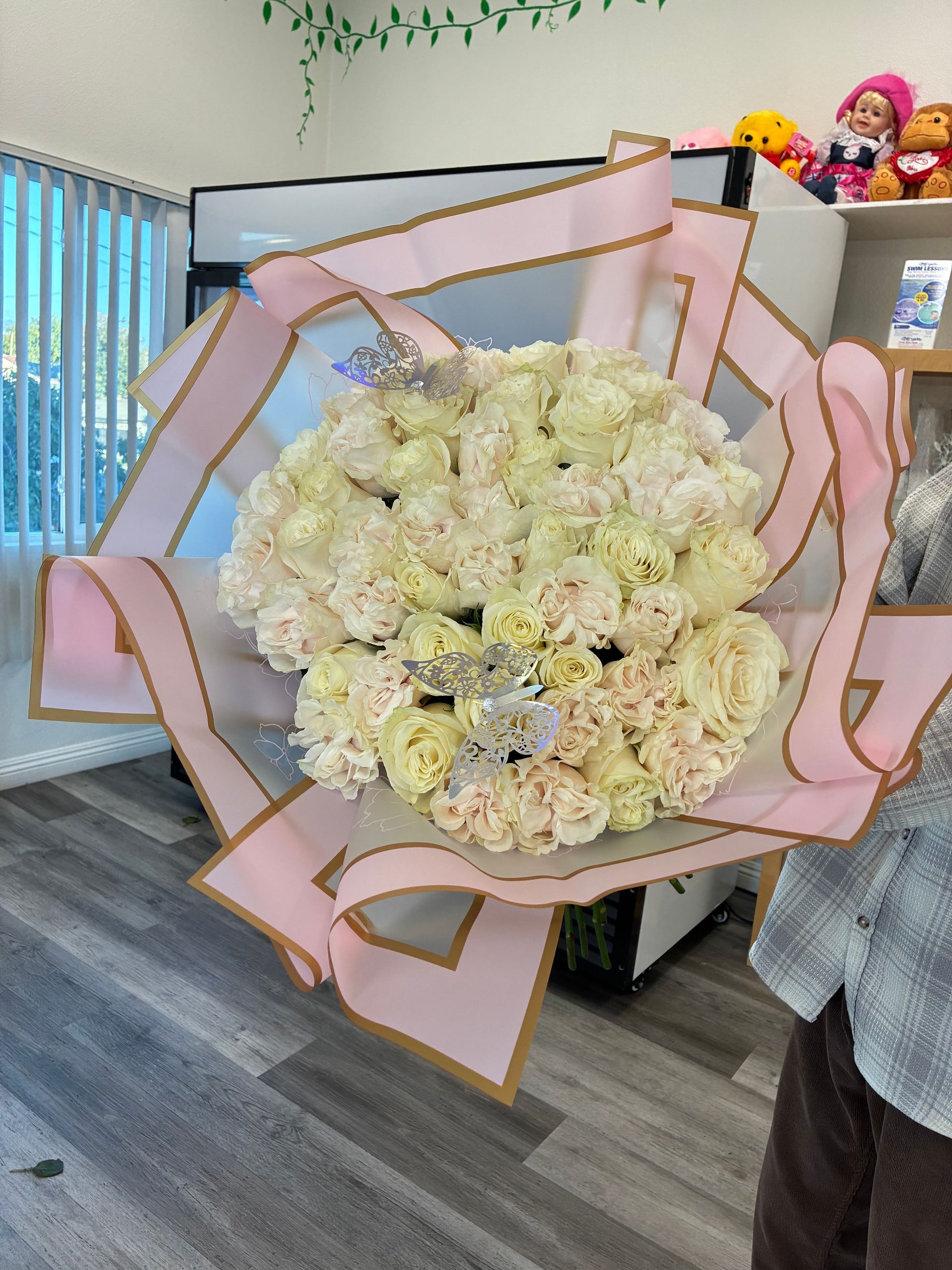A large bouquet of cream-colored roses wrapped in pink paper with gold edges, held indoors in a shop setting.