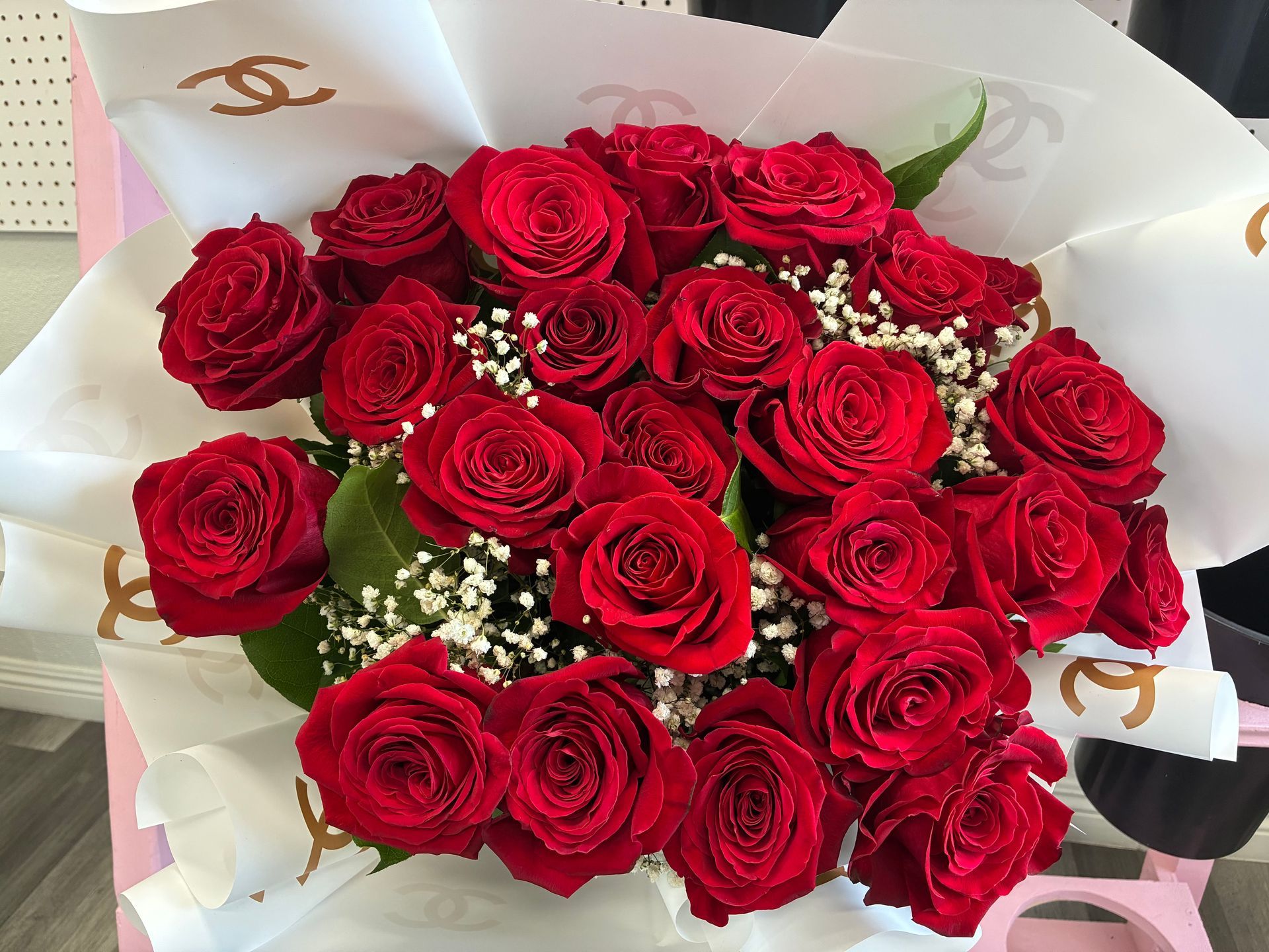 A bouquet of bright red roses accented with baby's breath, wrapped in white paper decorated with gold Chanel logos.