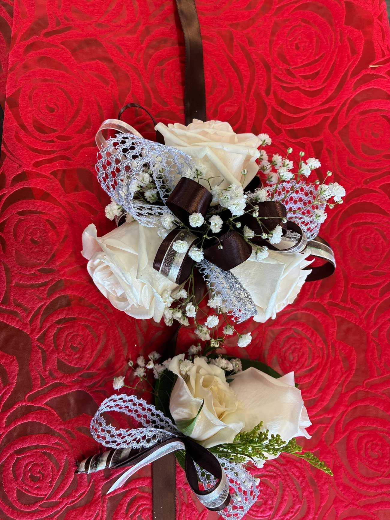 A white rose wrist corsage and matching boutonniere decorated with brown ribbons and silver lace on a red floral fabric.