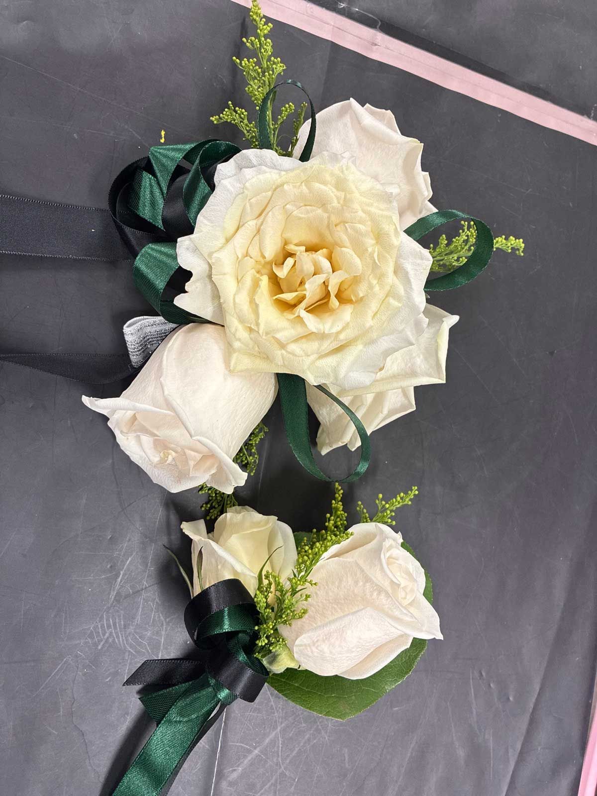 A white rose corsage and matching boutonniere featuring dark green ribbon accents and small sprigs of yellow greenery.