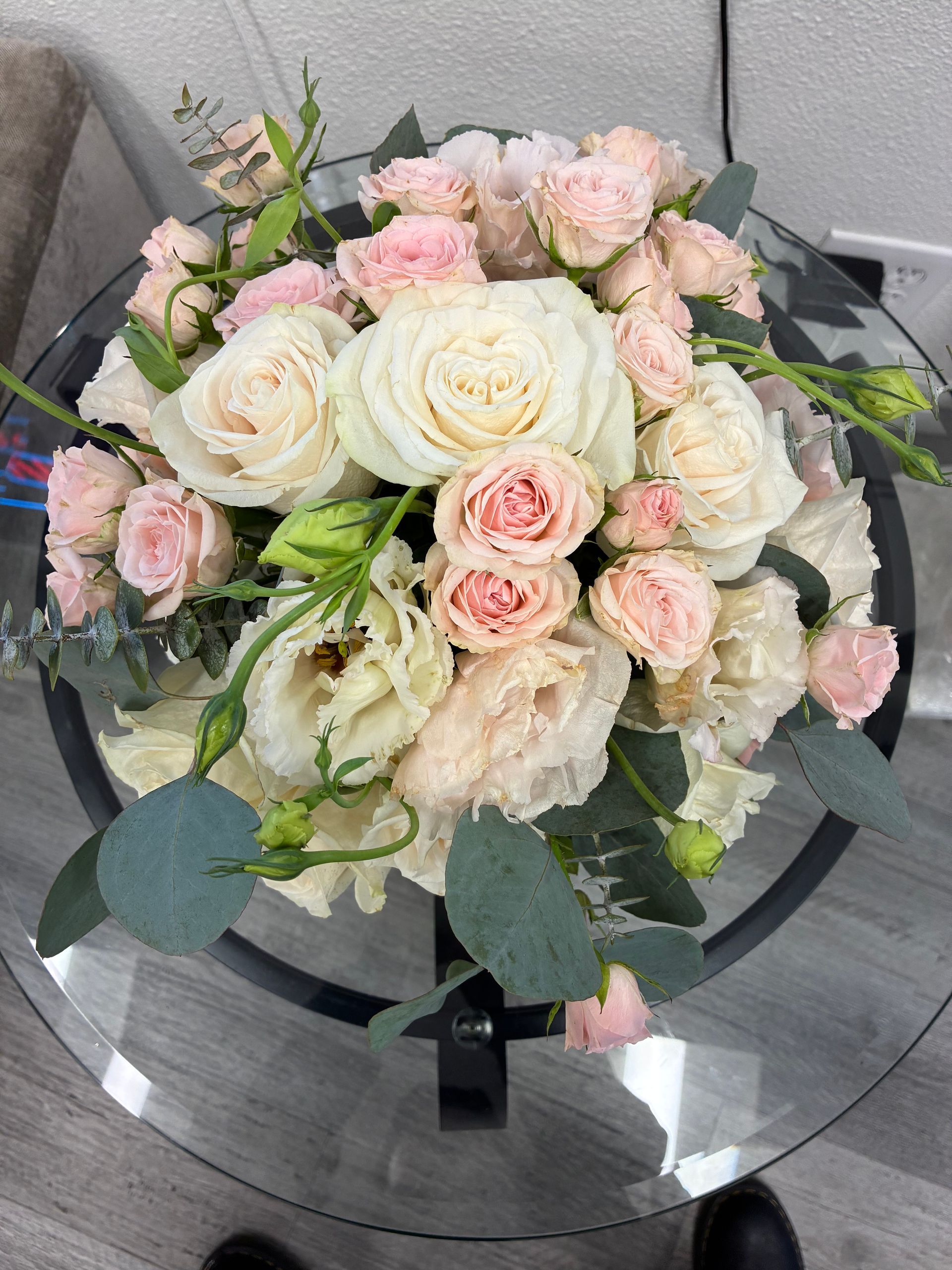 A lush bouquet of pale pink and white roses, eustoma, and eucalyptus greenery displayed on a glass-topped table.