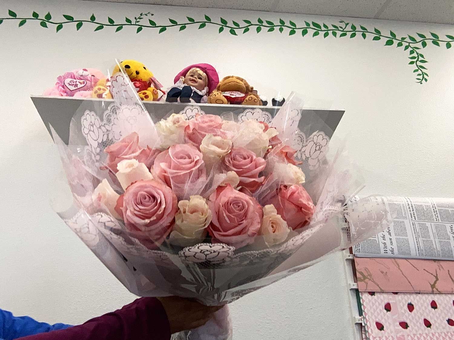 A bouquet of light pink and white roses wrapped in decorative paper, held up in front of a shelf with stuffed toys.