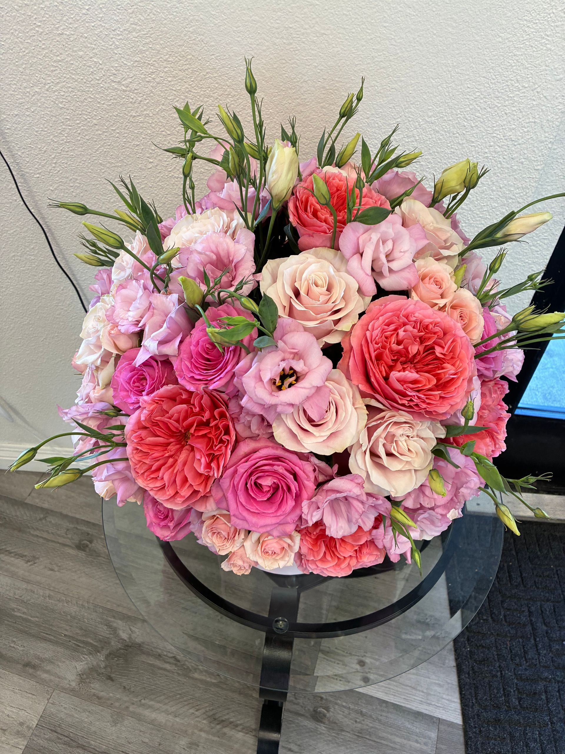 A lush, spherical bouquet of pink, coral, and peach roses and lisianthus in a white container on a glass-topped table.