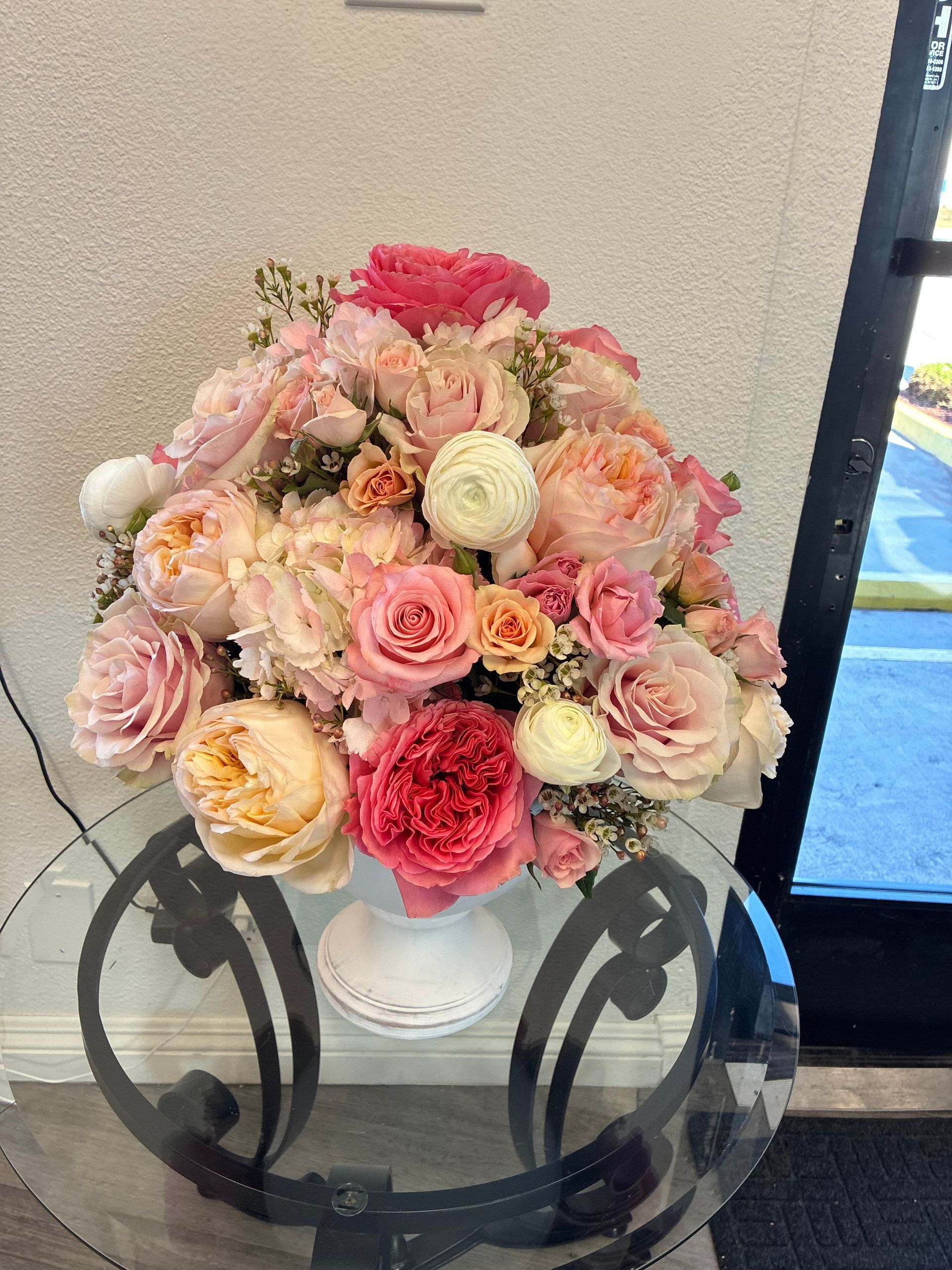 A lush, rounded floral arrangement of pink, peach, and white garden roses in a white pedestal vase on a glass table.