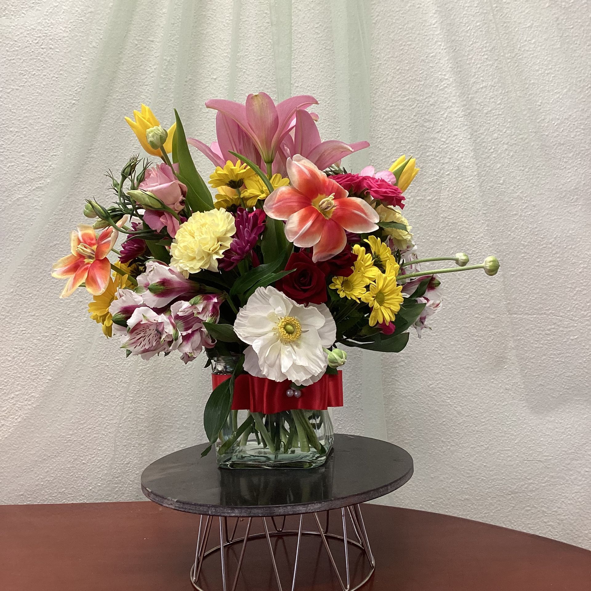 A colorful floral arrangement in a glass vase with a red ribbon, displayed on a circular black stand against a white wall.