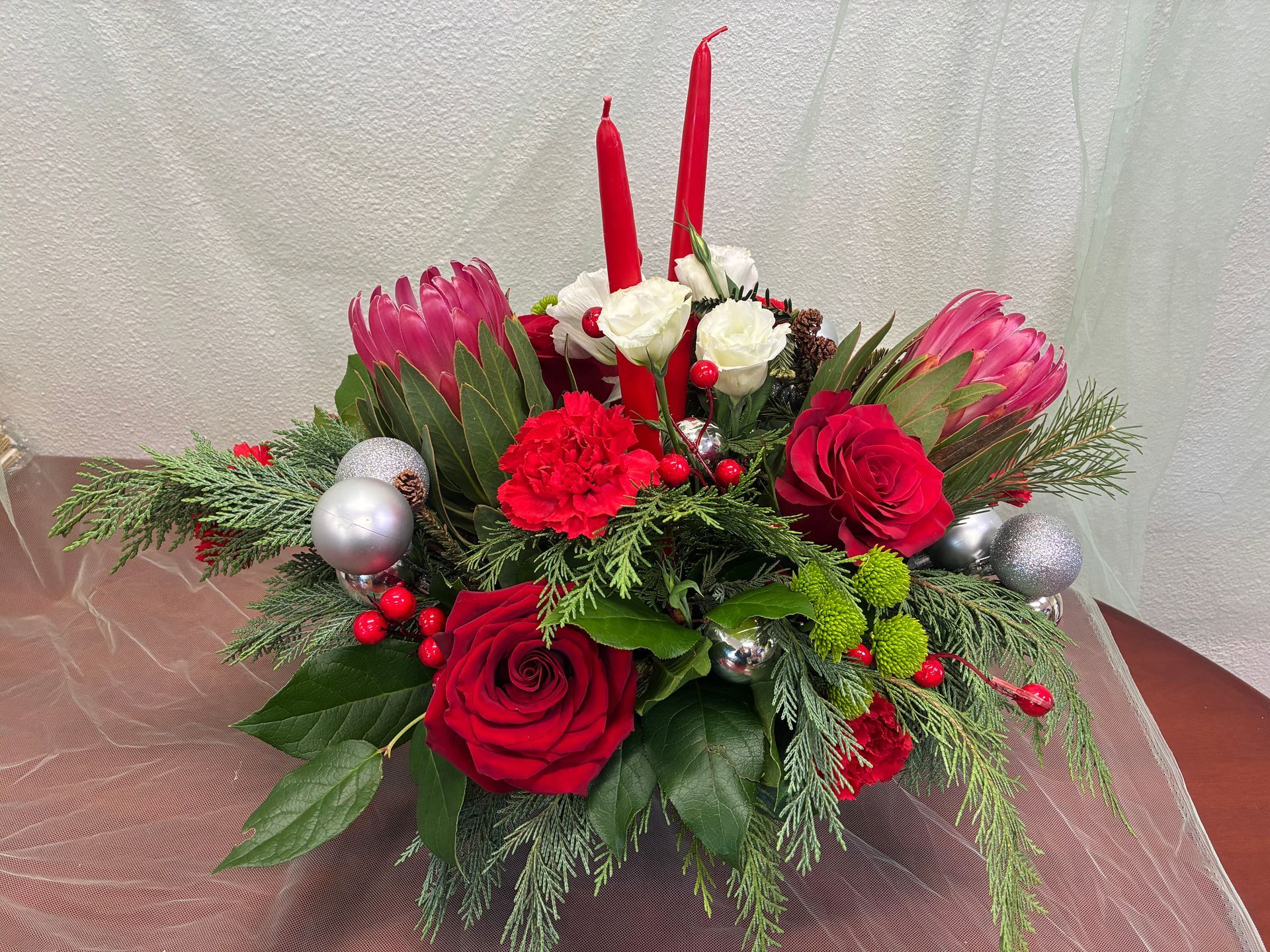 A Christmas floral arrangement with red roses, white roses, pink protea, greenery, silver ornaments, and two red candles.