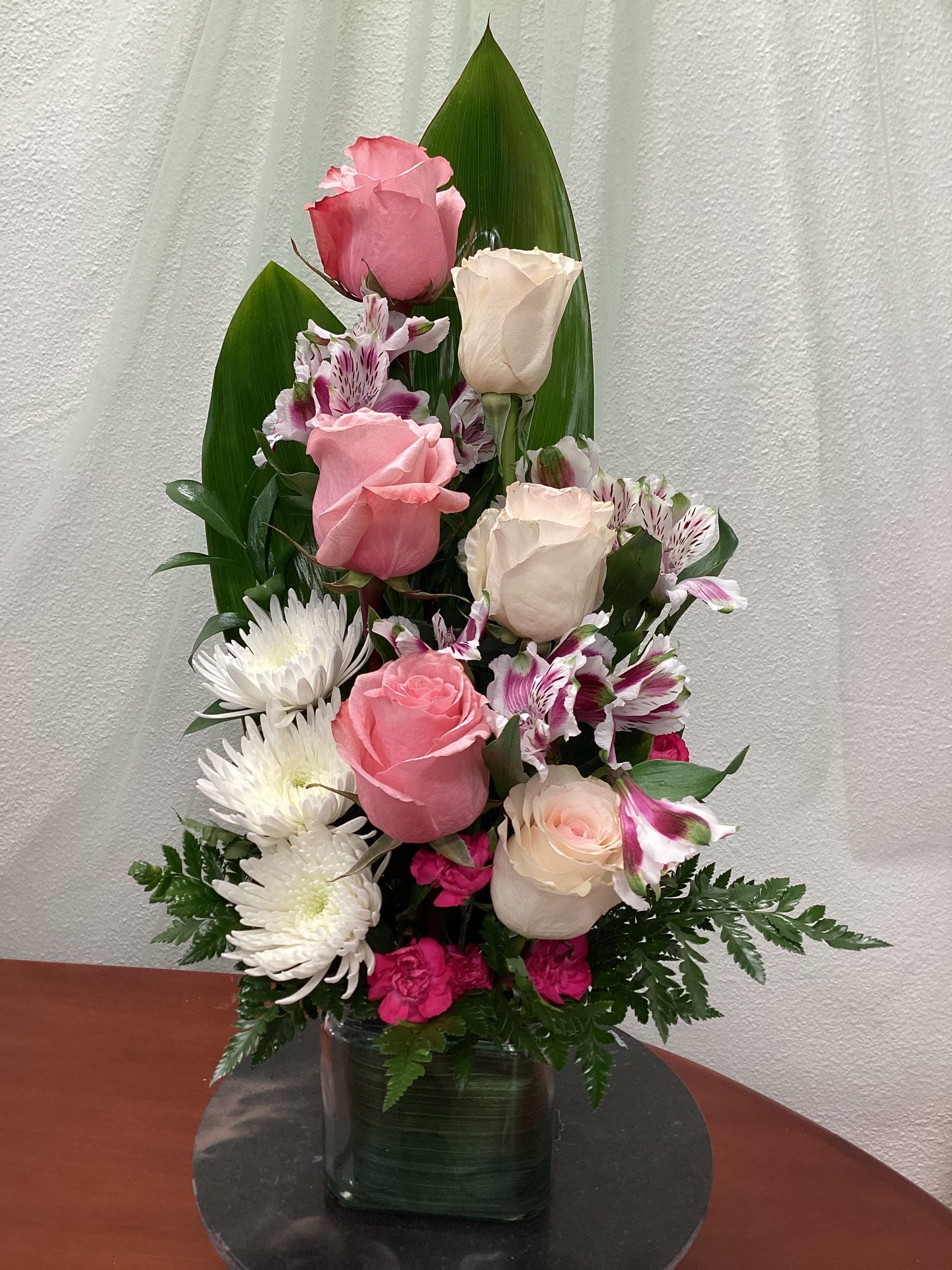 A vertical floral arrangement in a glass vase, featuring pink and cream roses, white chrysanthemums, and purple accents.