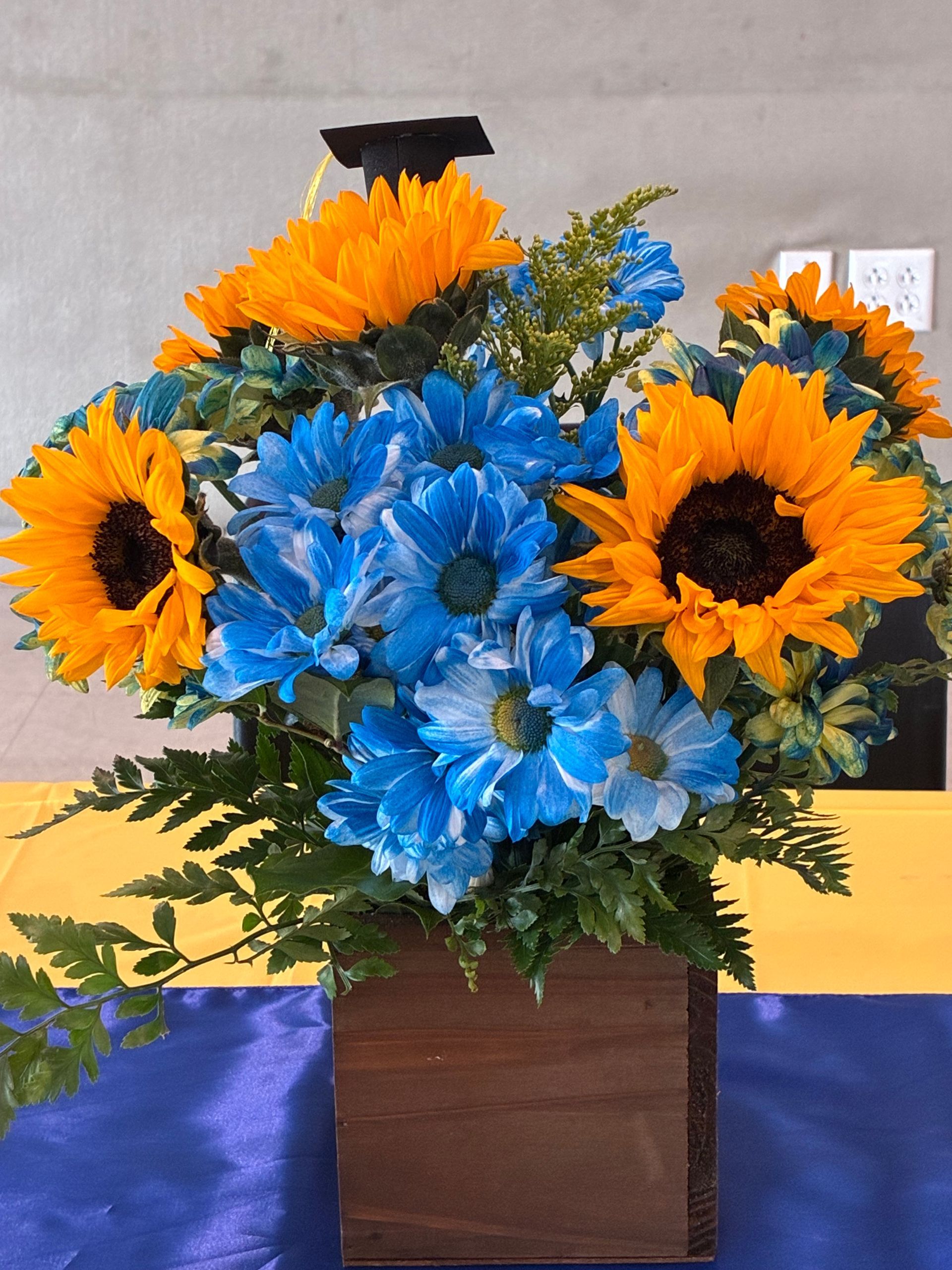 A graduation-themed floral arrangement with sunflowers and blue dyed chrysanthemums in a wooden box on a blue cloth.