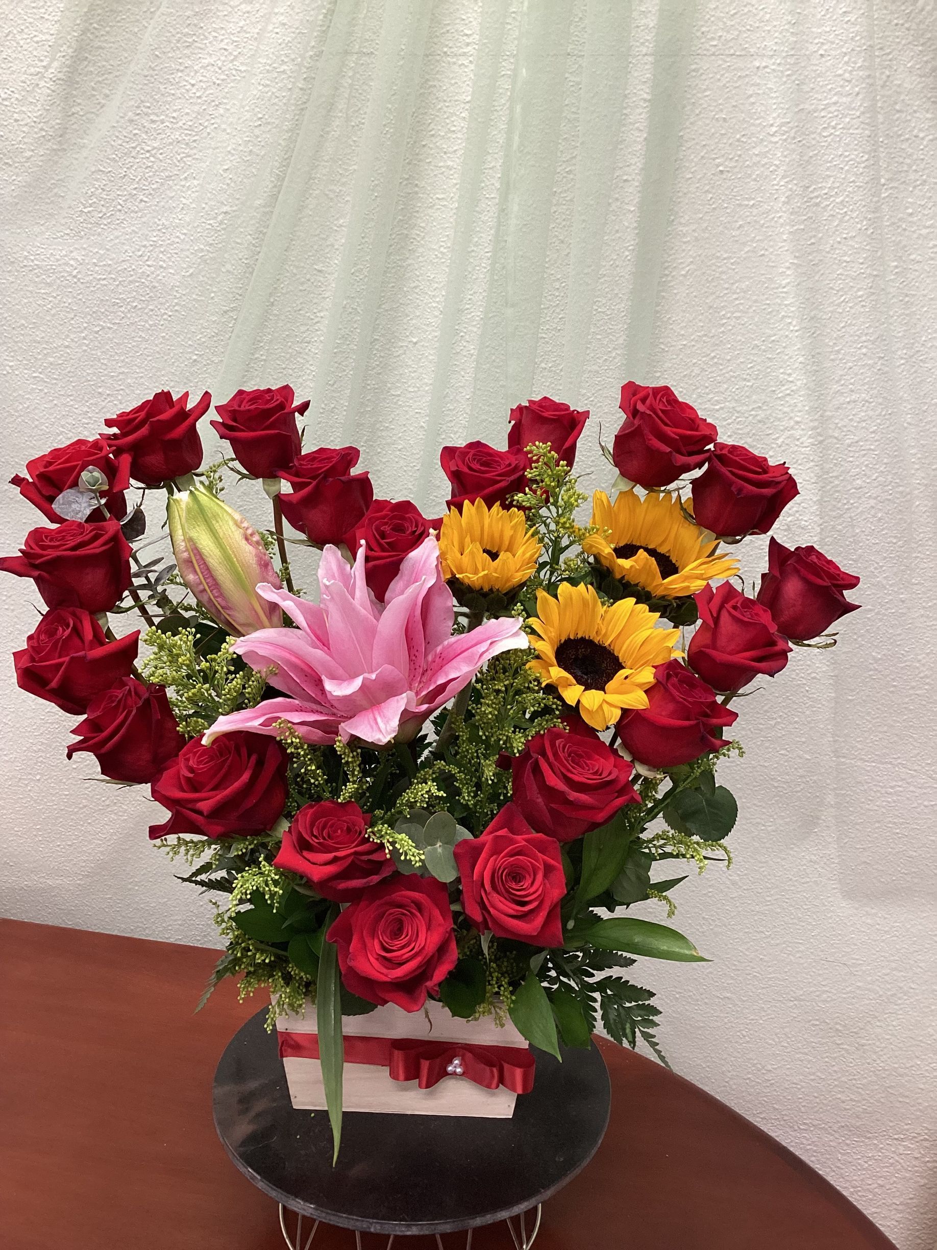 A heart-shaped floral arrangement of red roses, yellow sunflowers, and a large pink lily in a white box with a red ribbon.