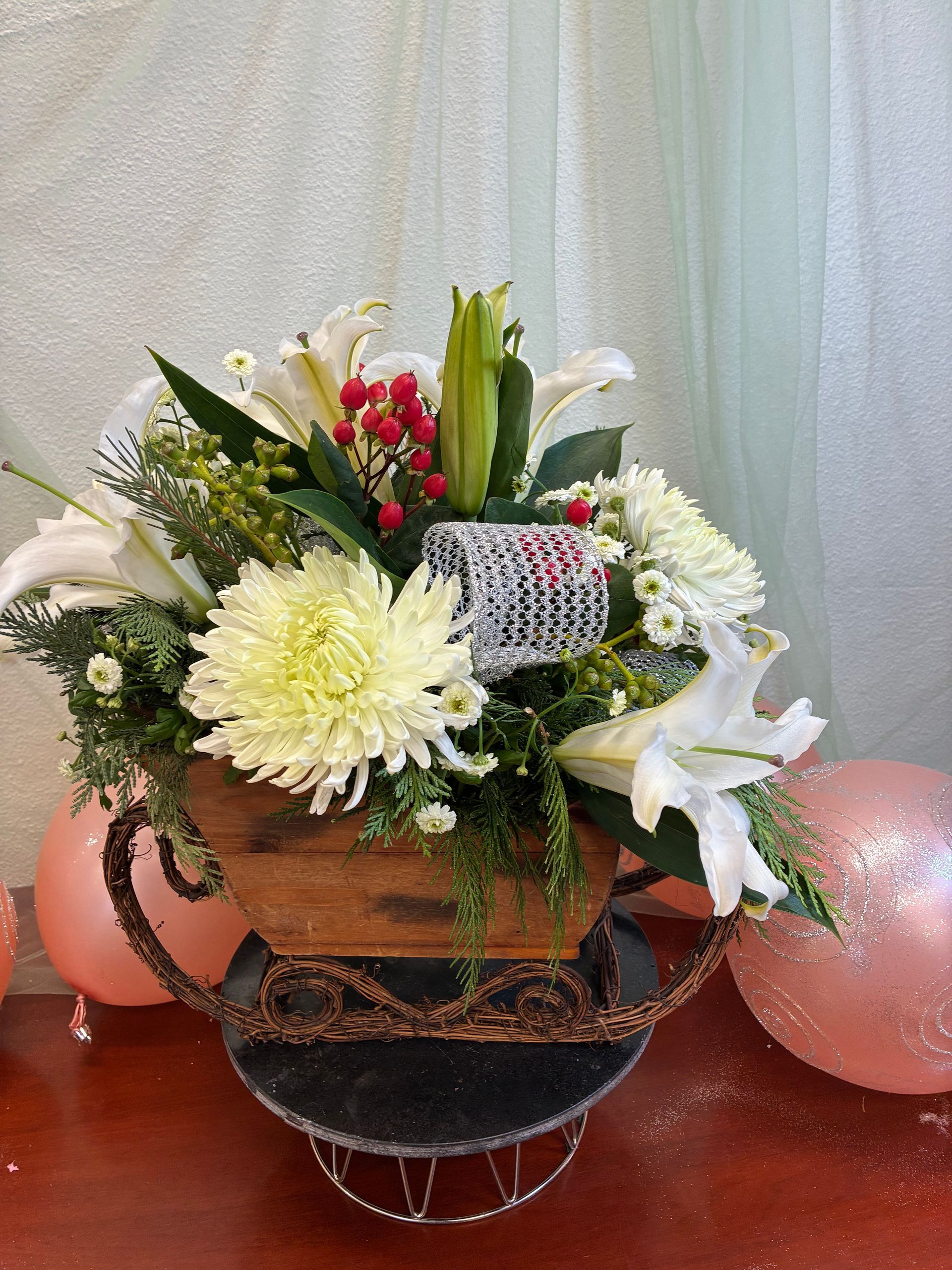 A holiday floral arrangement with white lilies, mums, and red berries in a rustic wooden sled-shaped planter.