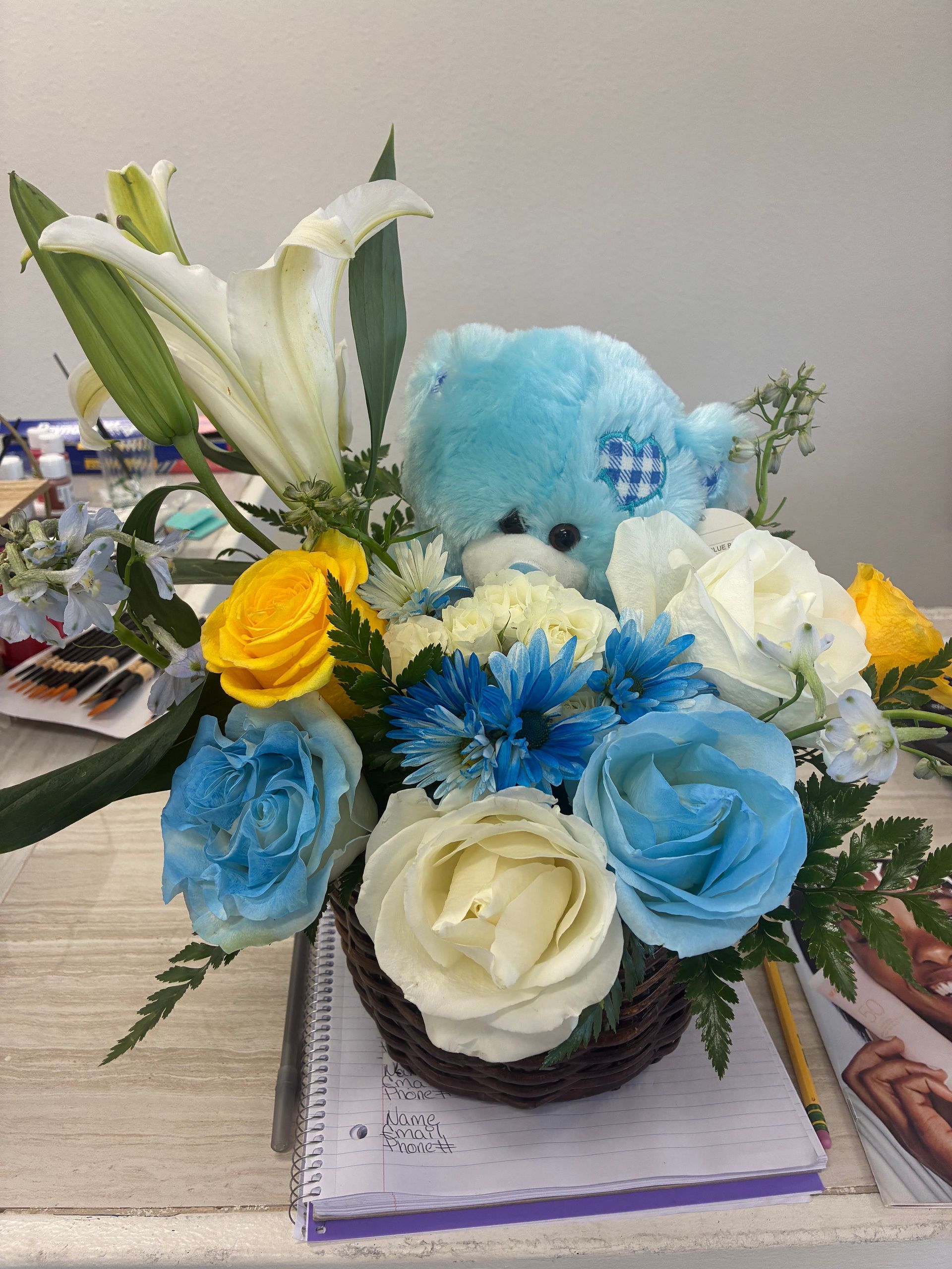 A floral arrangement in a basket featuring white lilies, blue and yellow roses, and a small blue teddy bear.