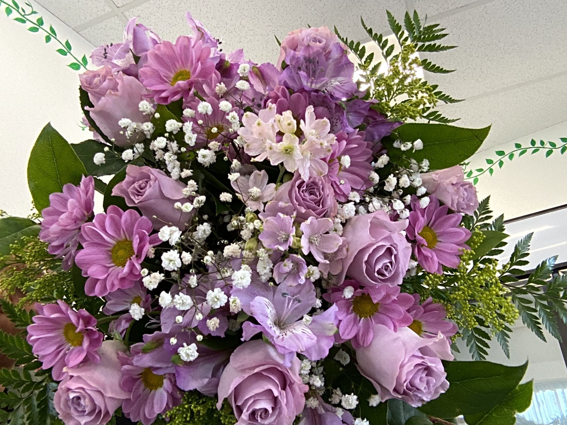 A bouquet of lavender and purple roses, daisies, and small white flowers with green foliage against a white background.