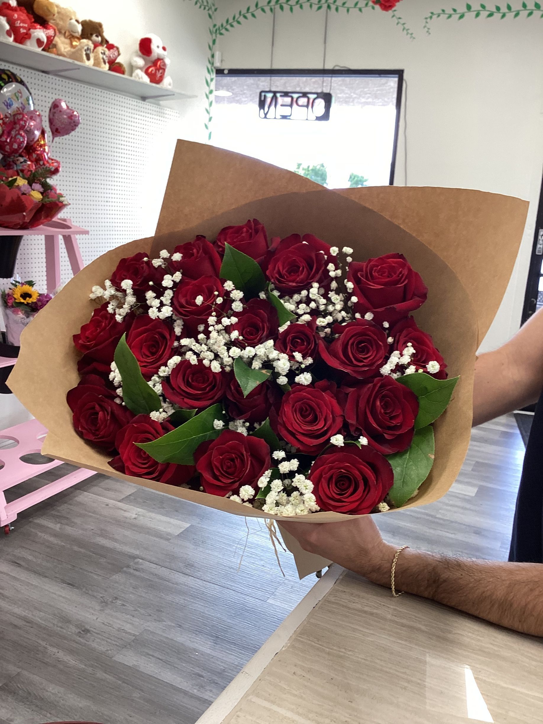 A bouquet of red roses with baby’s breath, wrapped in brown paper, held in a person's hands inside a floral shop.