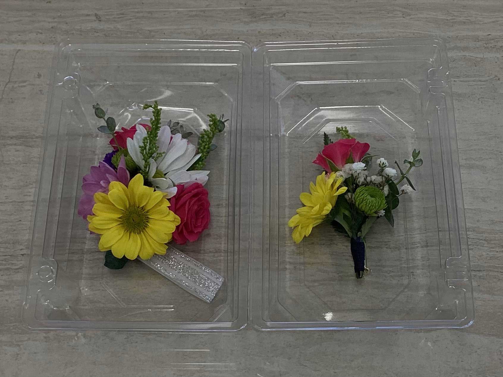 Two floral arrangements in clear plastic containers, one a corsage with a glittery band and the other a boutonnière.