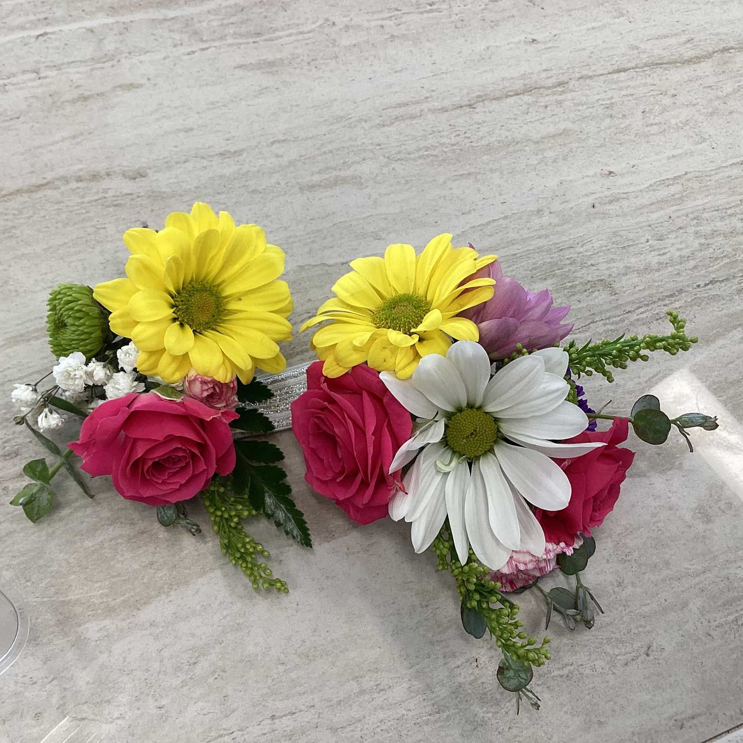 Two floral boutonnieres featuring yellow and white daisies, pink roses, and green filler on a light marble surface.