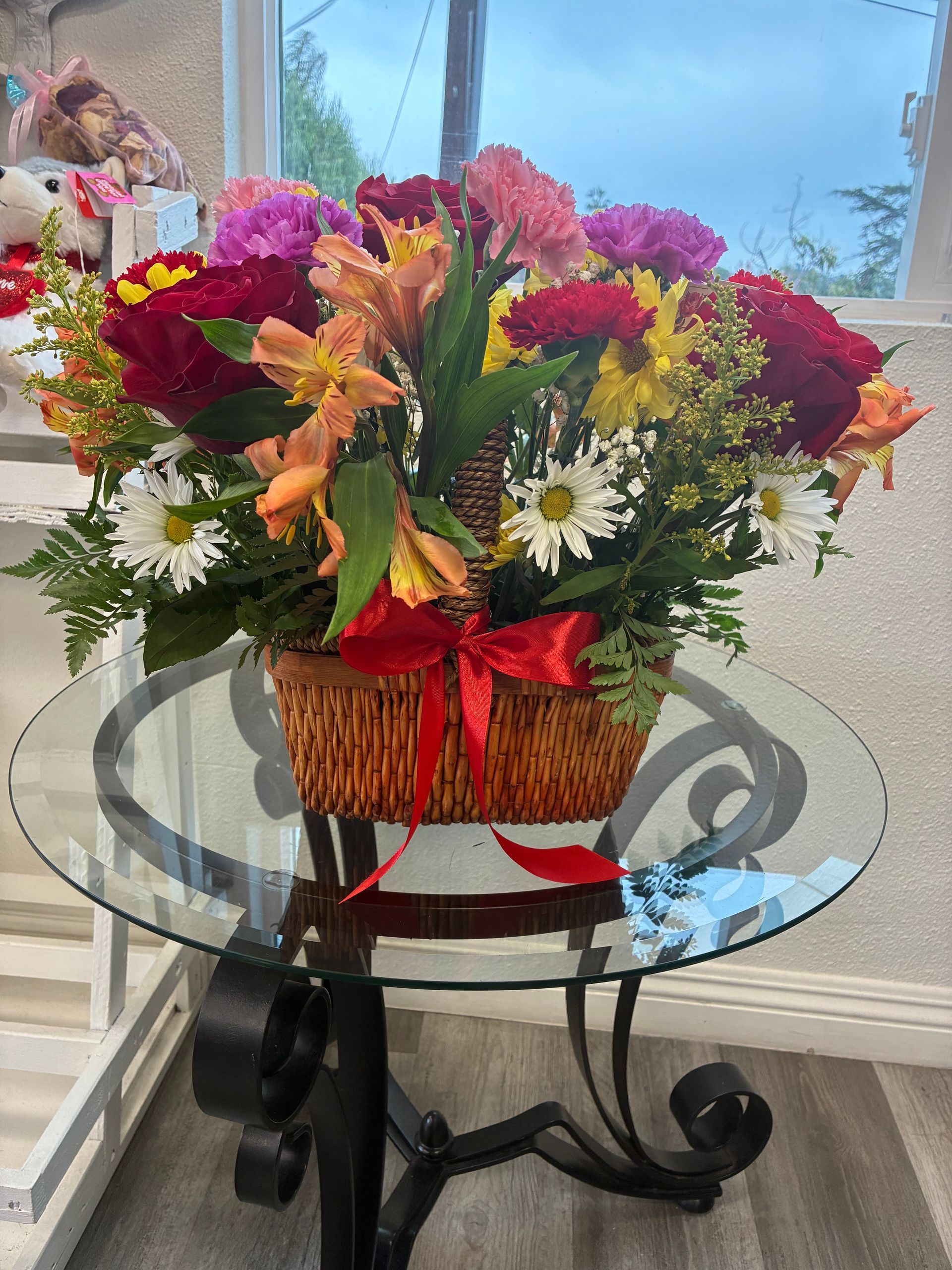 A colorful floral arrangement in a woven basket with a red ribbon, sitting on a round glass-top metal table.