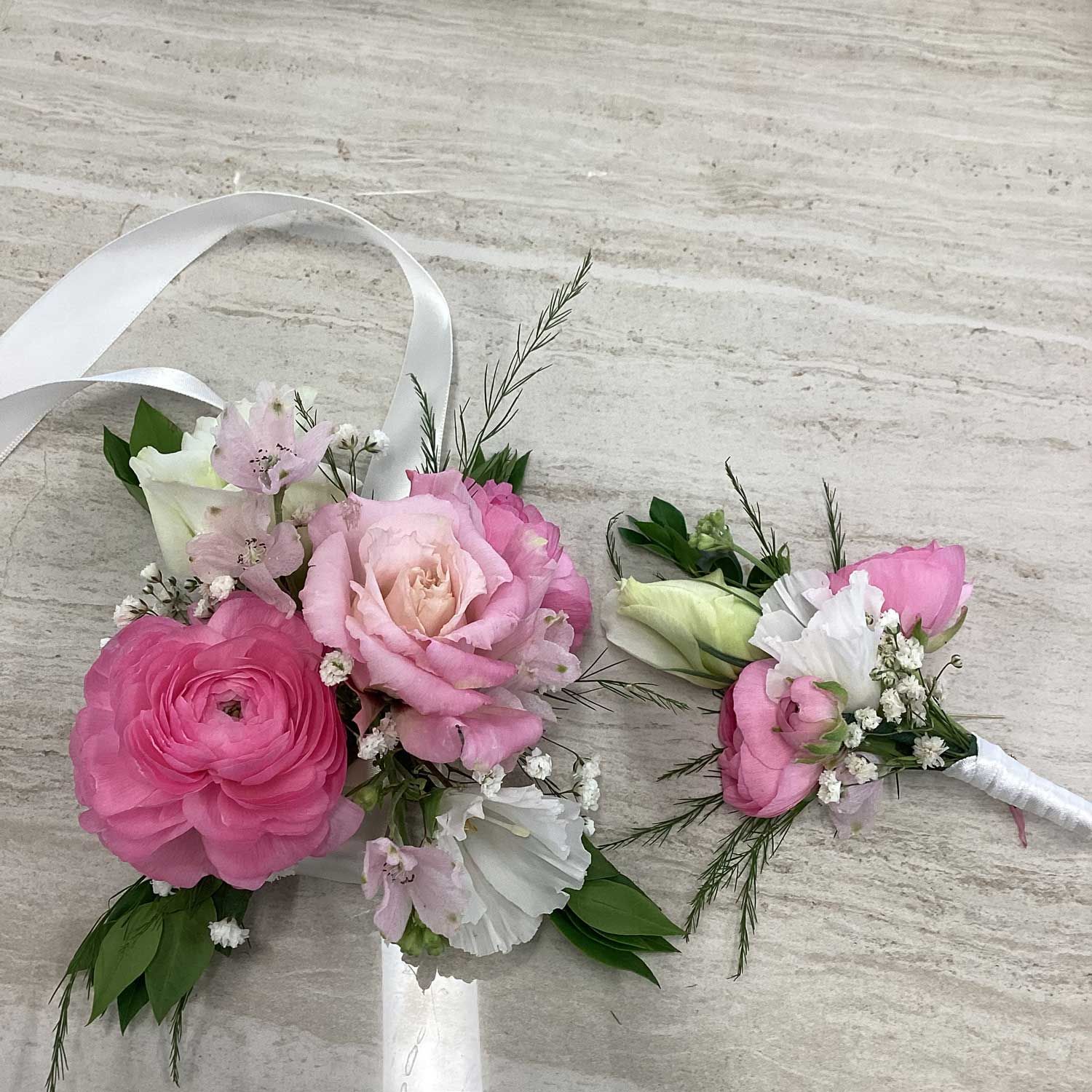 A floral wedding wrist corsage and a matching boutonniere featuring pink roses, white lisianthus, and baby’s breath.