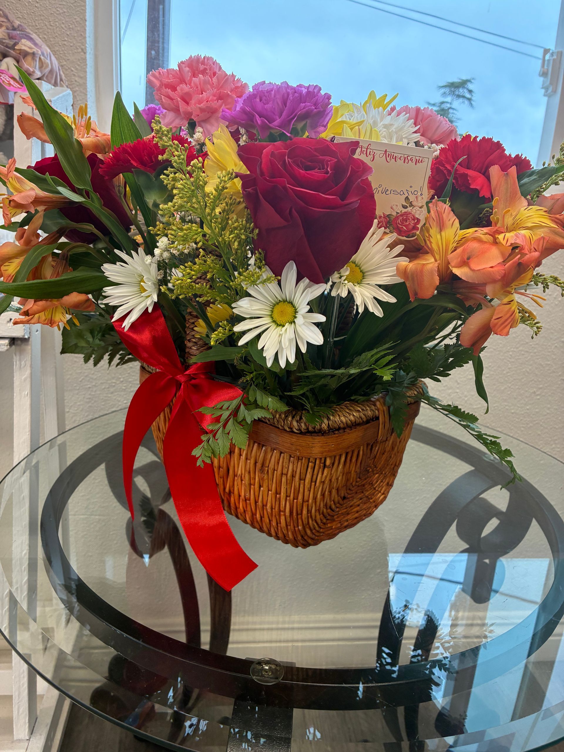A wicker basket filled with colorful flowers including a large red rose and daisies, accented by a red ribbon on a table.
