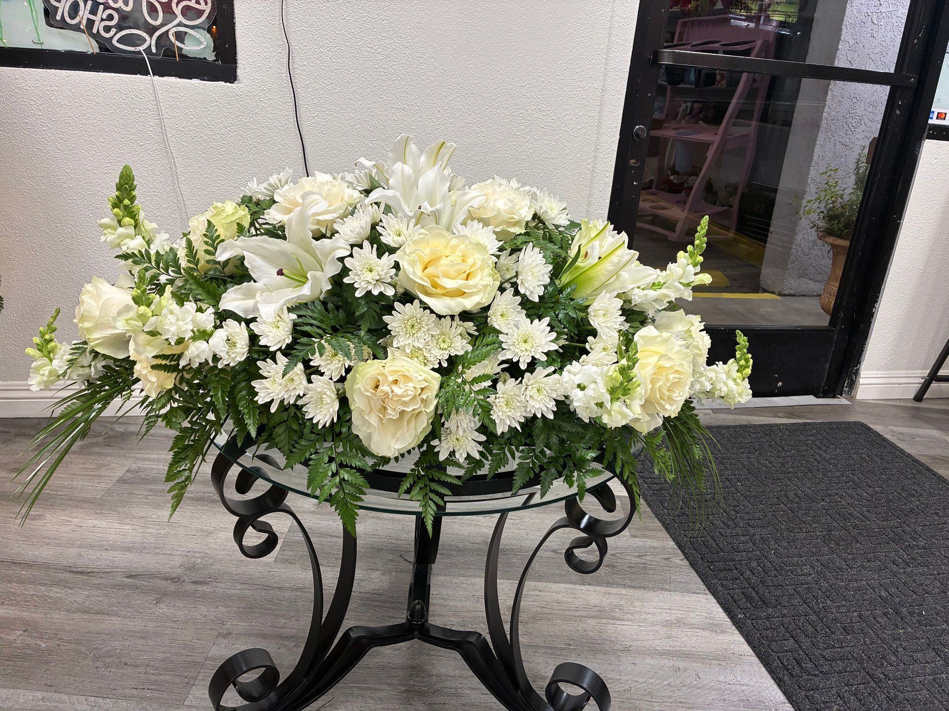 A large arrangement of white lilies, roses, and carnations sits on a black metal table in an indoor setting.