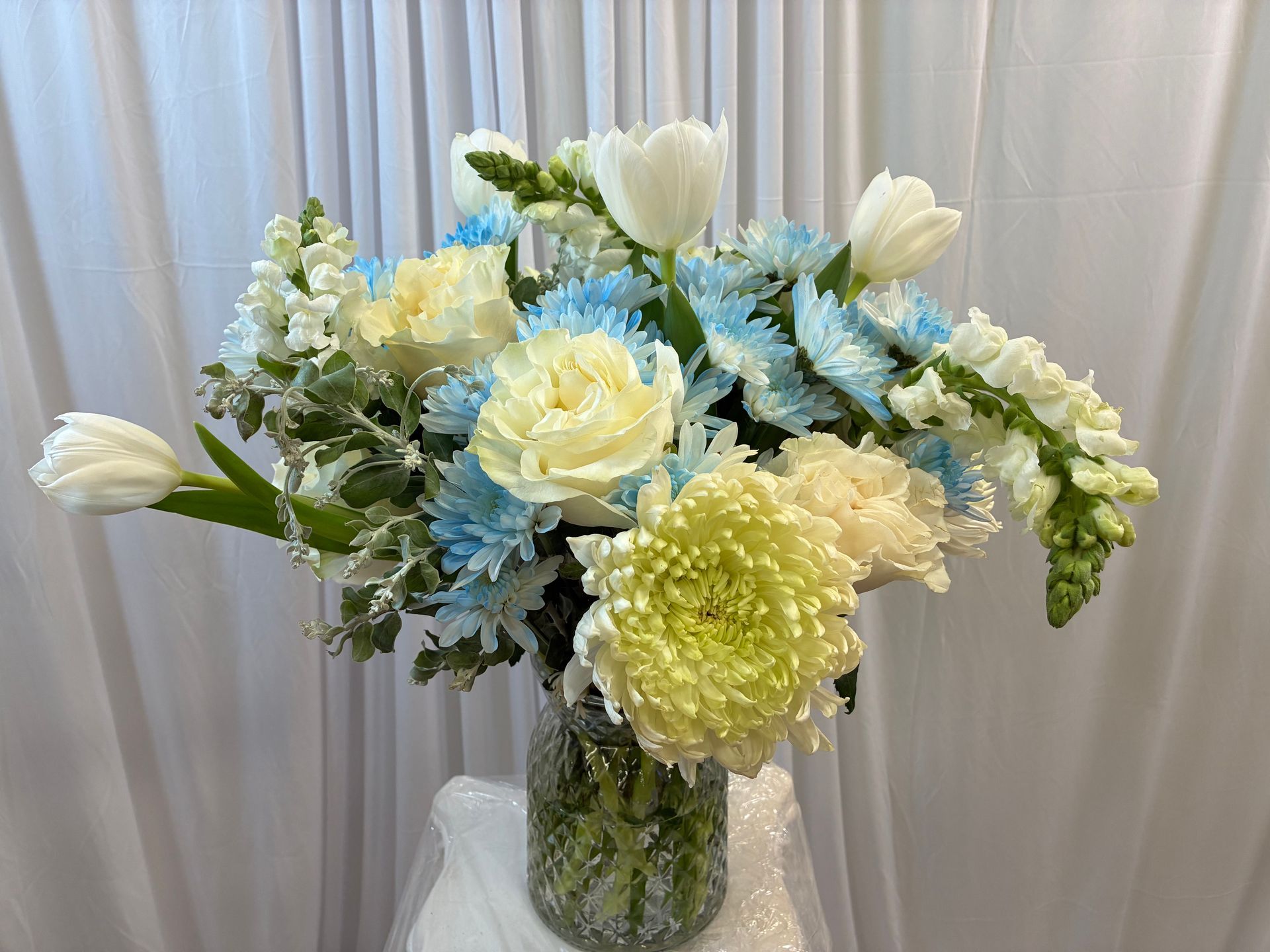 A floral arrangement with white tulips, light blue chrysanthemums, and snapdragons in a textured glass vase.