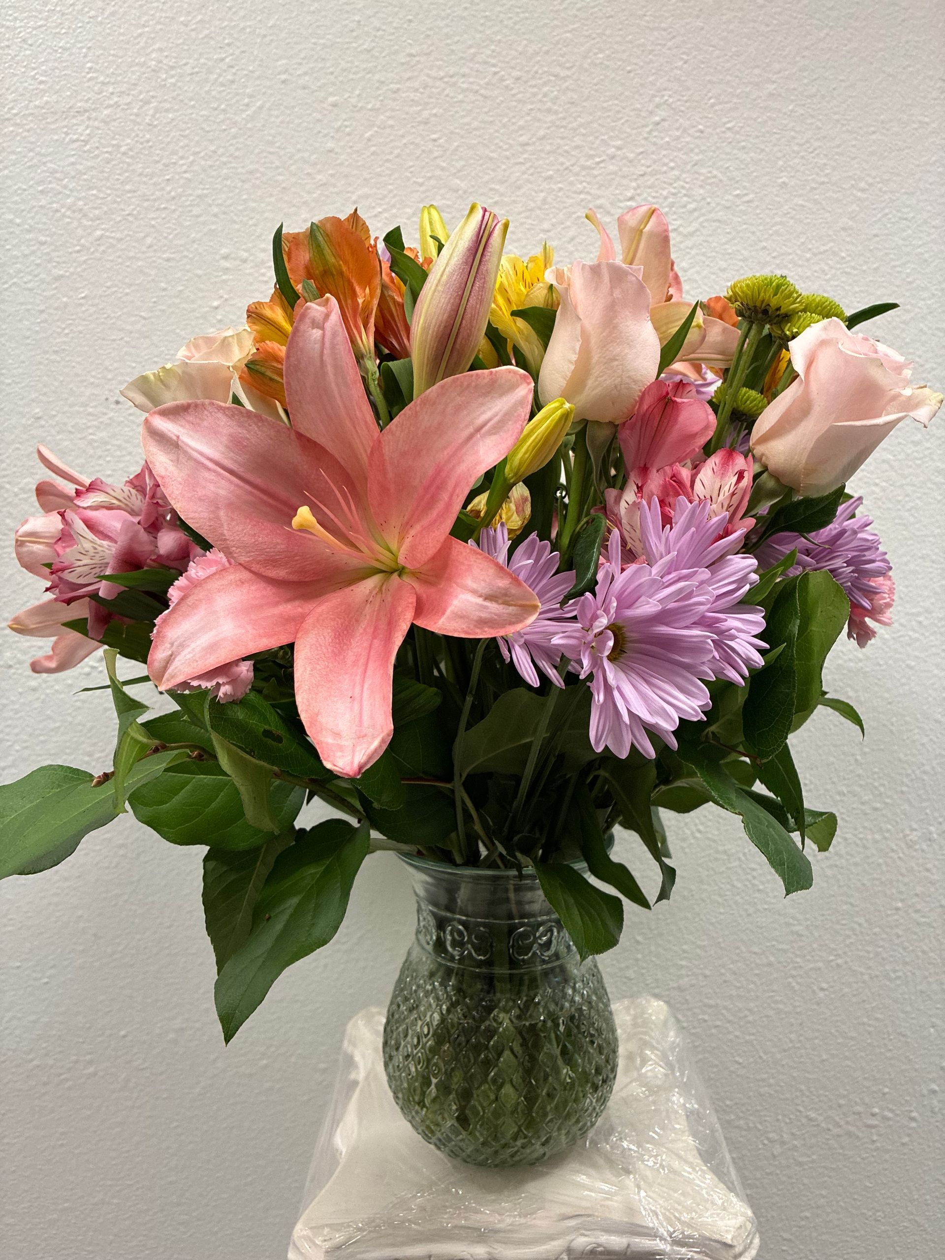 A colorful bouquet of pink lilies, soft pink roses, and purple chrysanthemums arranged in a clear glass vase.
