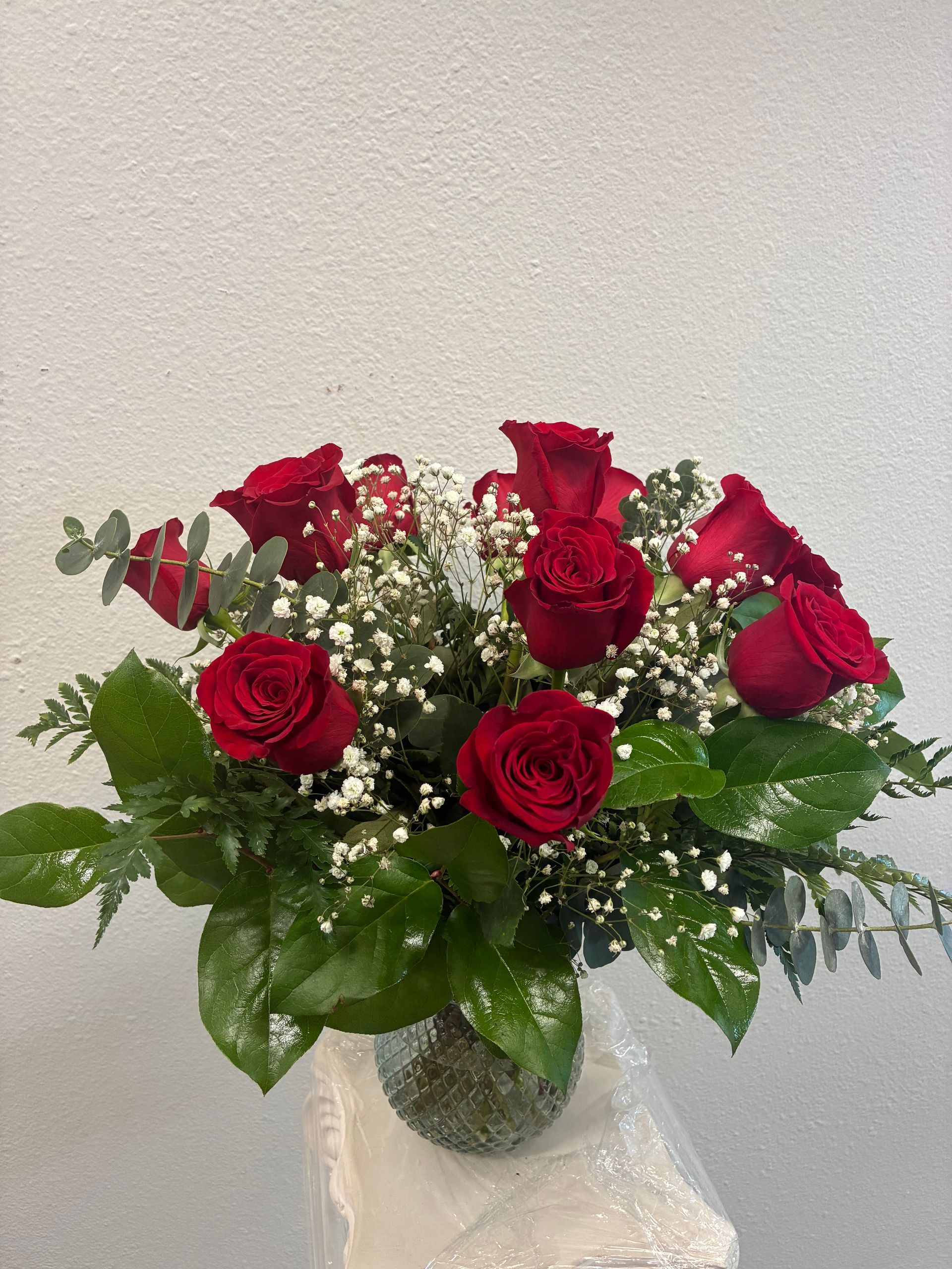 A bouquet of deep red roses and white baby's breath with lush green foliage in a textured glass vase.
