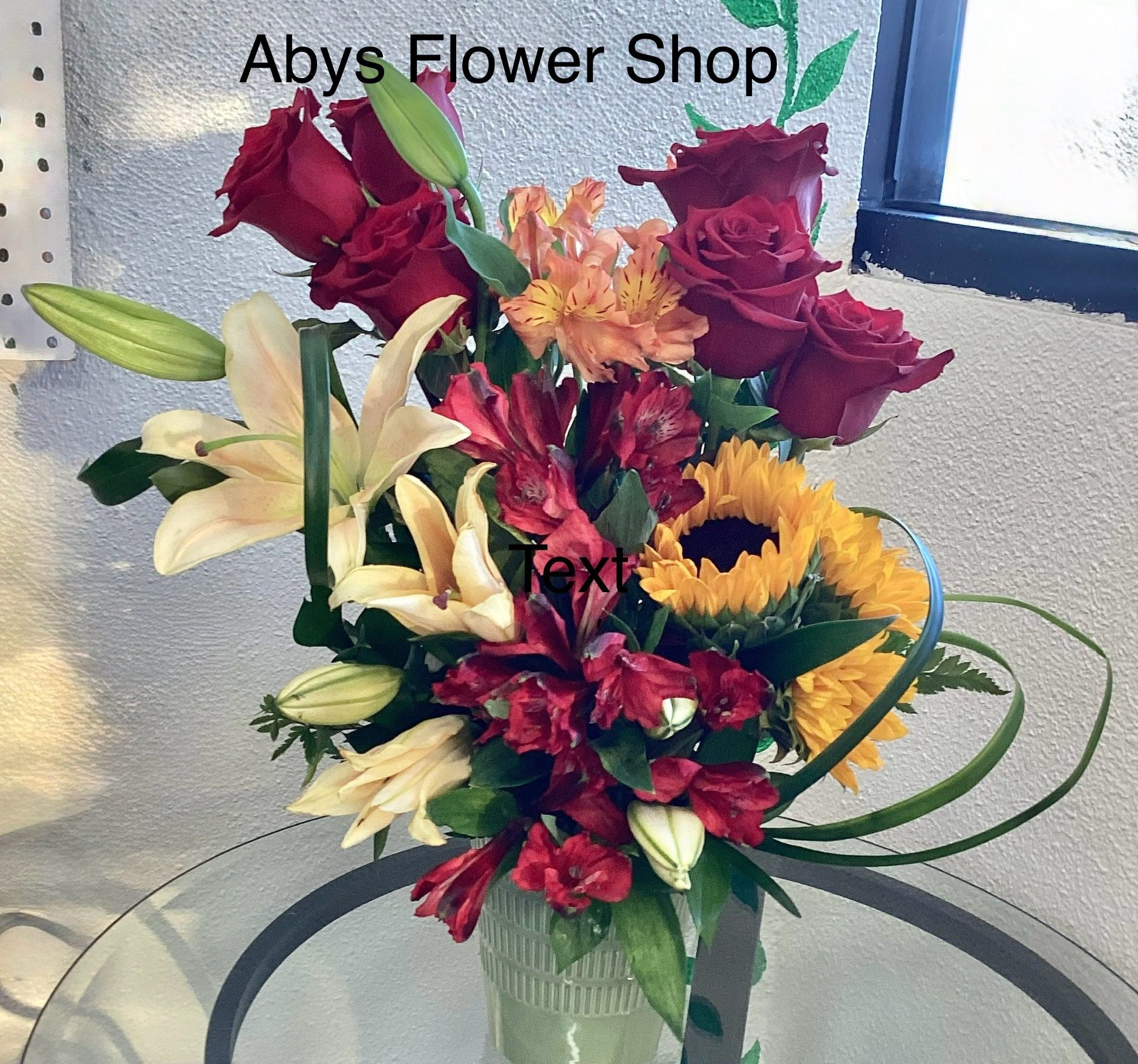 A vibrant bouquet featuring red roses, yellow lilies, a sunflower, and dark pink alstroemeria in a glass vase.