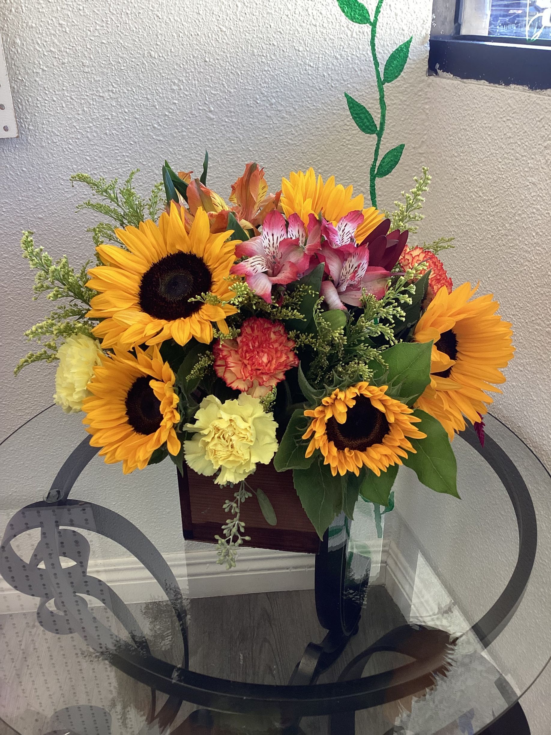 A colorful floral arrangement featuring sunflowers, pink alstroemeria, and yellow carnations in a vase on a glass table.
