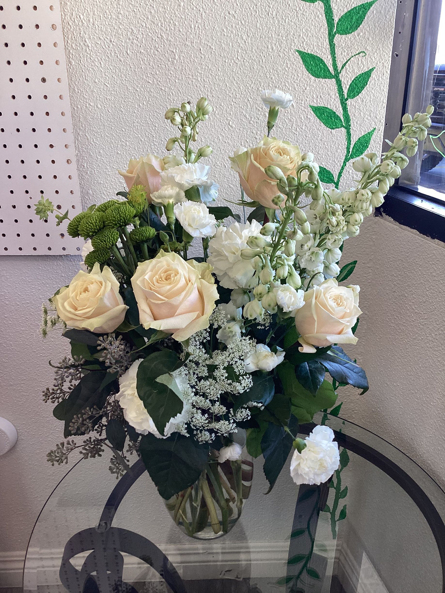 A bouquet of white and pale peach roses, snapdragons, and greenery in a glass vase sitting on a glass table.