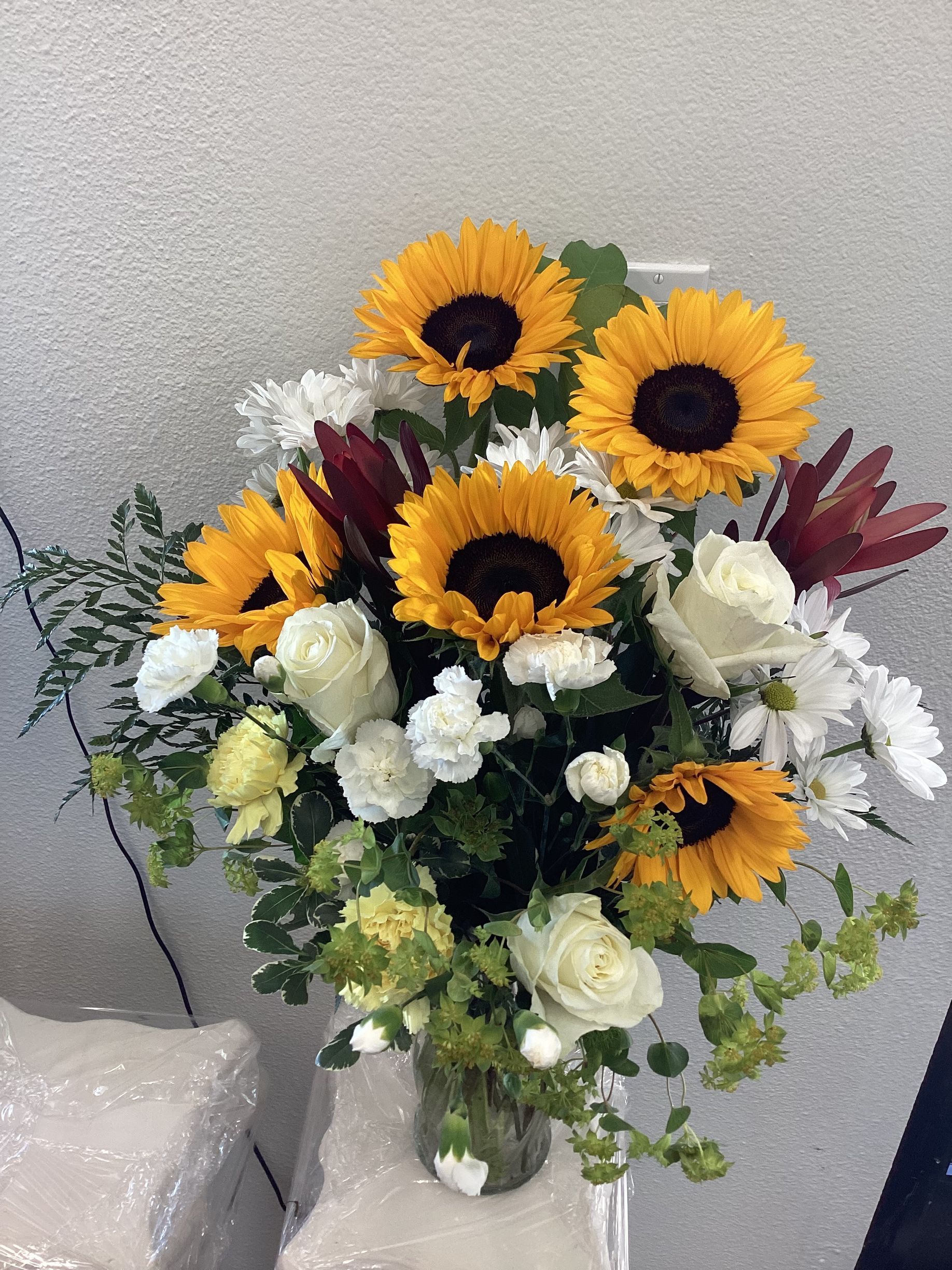 A vibrant floral arrangement featuring bright yellow sunflowers, white roses, and dark purple blooms in a clear glass vase.